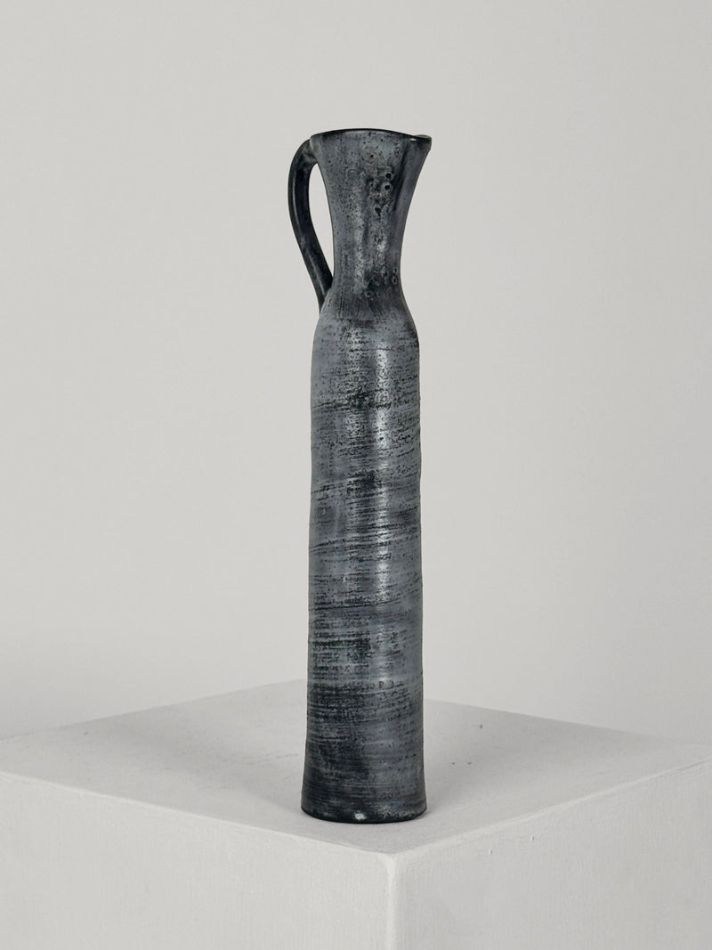 A tall, slender, and textured black Ceramic Bottle by Jacques Pouchain from Figuresdesign, featuring a small handle and displayed on a white pedestal against a plain gray background. The bottle showcases its handmade, artisanal qualities with a narrow neck and widening base, reminiscent of Pouchain's signature style.