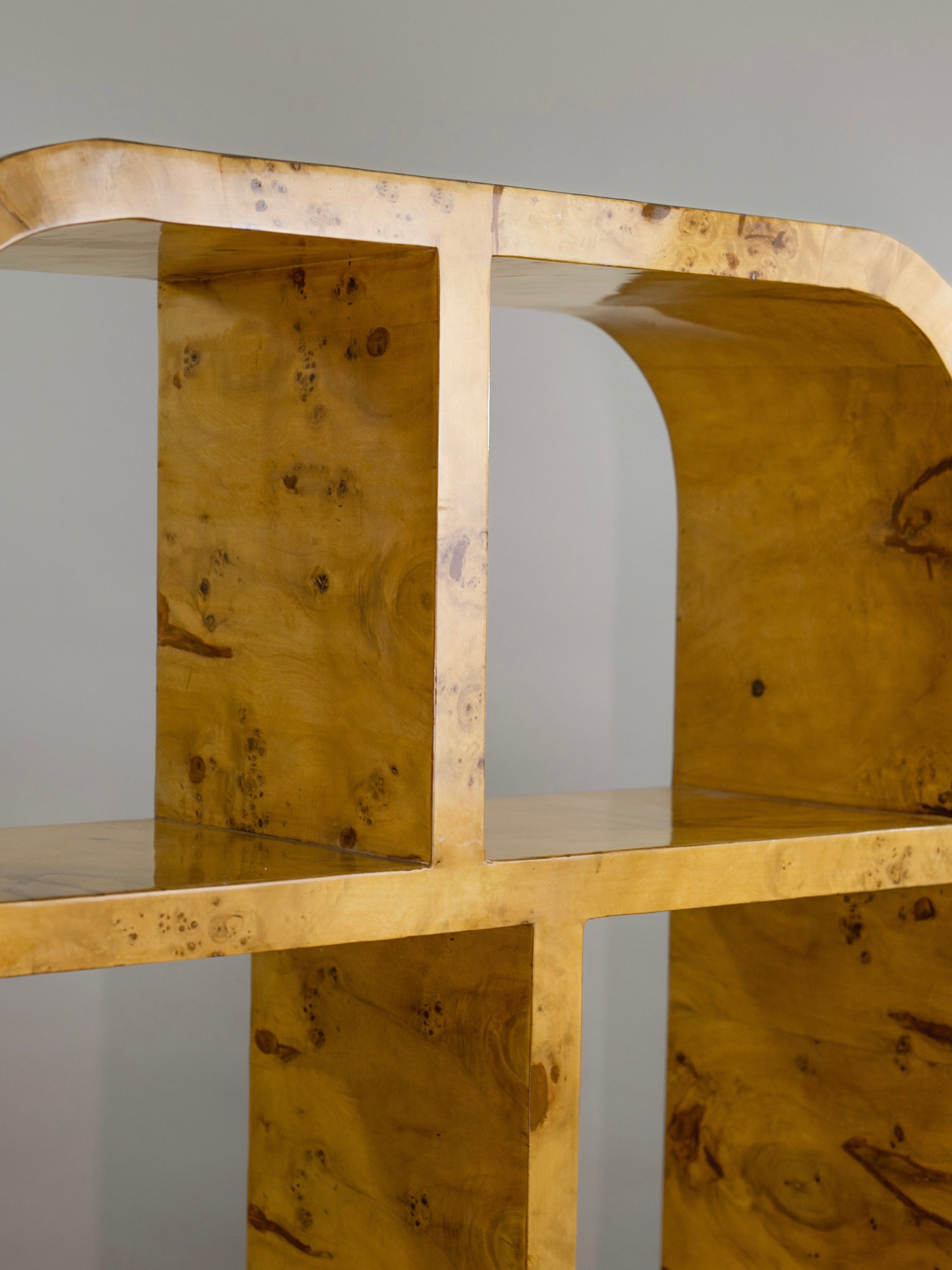 The 'Calisto' Double Bookcase by Meola Interiors features a wooden shelf with a glossy marbled finish, rounded edges, and geometric design in mid-century style. Its light brown wood showcases natural grain patterns, enhancing its minimalist look against a muted gray background.