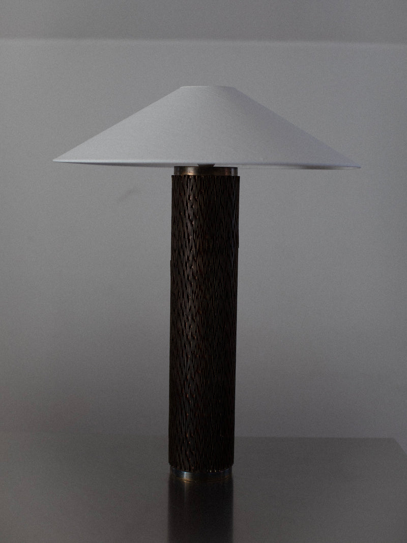 A modern Brutalist Floor Lamp by Out For Lunch, featuring a cylindrical dark-textured base and a wide conical white lampshade, is positioned on a metallic surface against a plain grey background. The lamp combines contemporary design elements with a minimalist aesthetic.