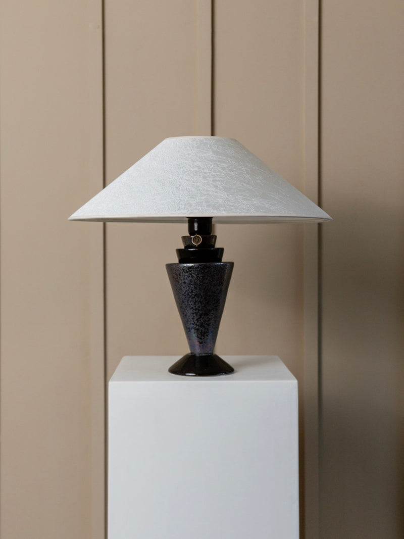The Bottega Jacobs Vintage Ceramic Table Lamp with a Silk Shade from the 1970s, designed in the Hollywood Regency style, features a textured white silk lampshade and a dark, cone-shaped ceramic base. Expertly handmade in Italy, it is displayed gracefully on a white pedestal against vertical beige panels.