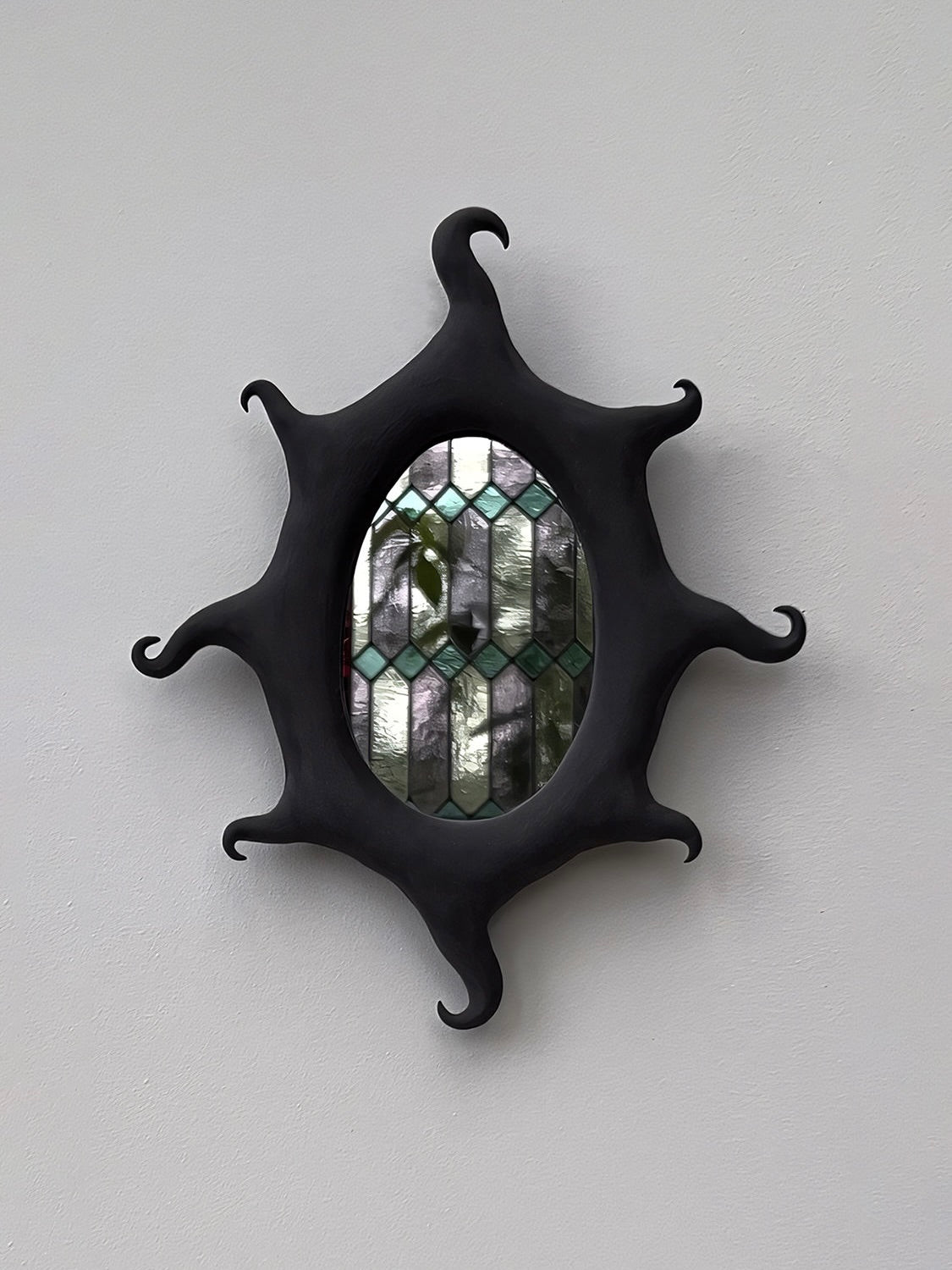 Mounted on a plain gray wall, the Black Sun Mirror by Solenne Belloir exudes a magical presence with its black, wavy-edged frame and stained glass design. The green and clear geometric patterns shimmer beautifully under the light, their elegance amplified by a lustrous black matte finish.