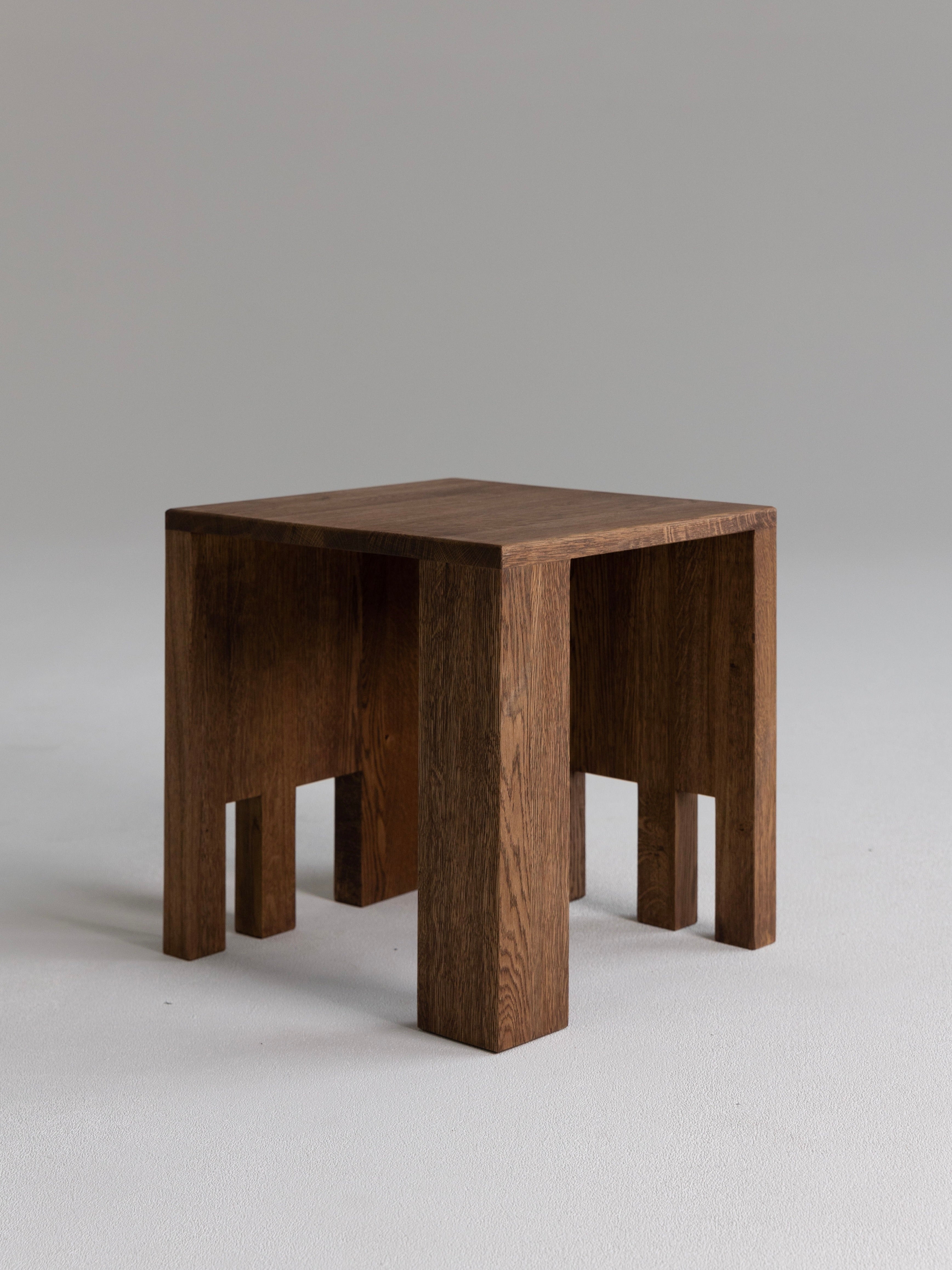 The Good Living&Co.'s "Dente" Oak Wood Stool features a square seat and four blocky legs on a light grey background. Its visible wood grain highlights its rustic, handmade appearance, reflecting exceptional craftsmanship and minimalist design.