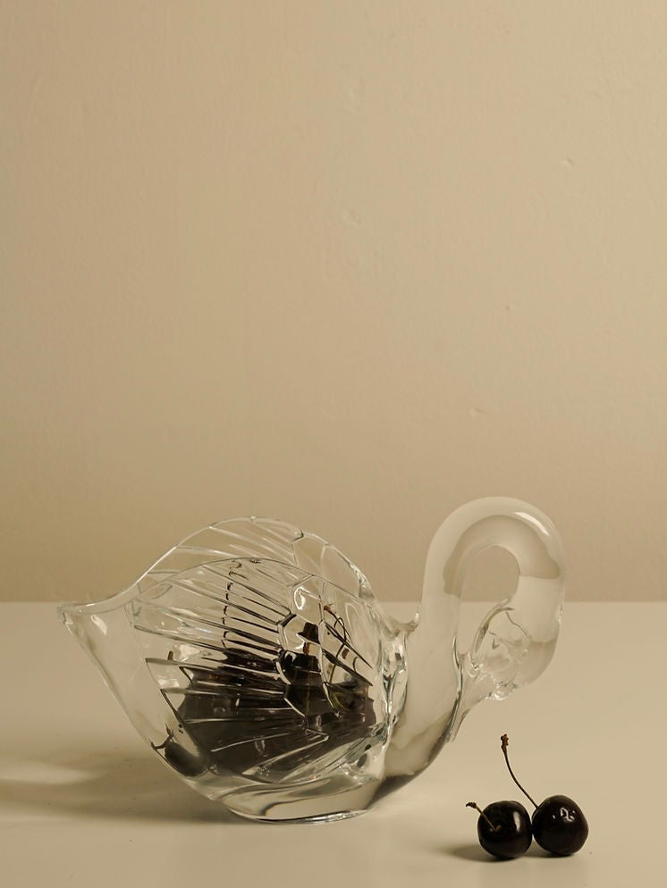 A clear glass Swan Bowl from Les Objets Amsterdam, known for its intricate detailing, is placed on a light-colored surface. Next to the bowl are two dark cherries resting on the same surface. The background is a plain beige wall.