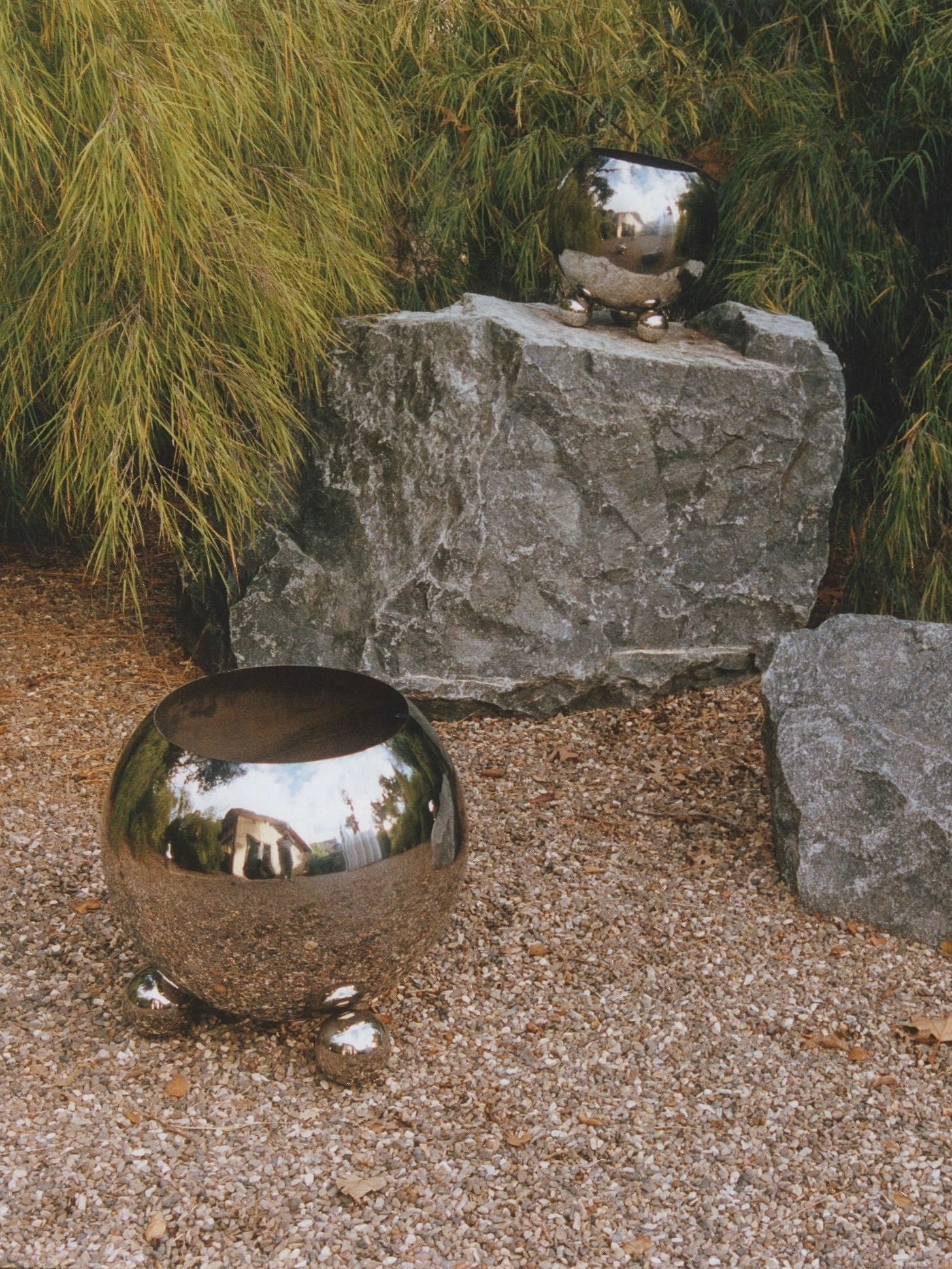 A garden scene featuring a large MUHLY Selene Planter on a gravel path, and a smaller similar planter placed on a large rock in the background. The serene setting is completed by green foliage forming the backdrop.