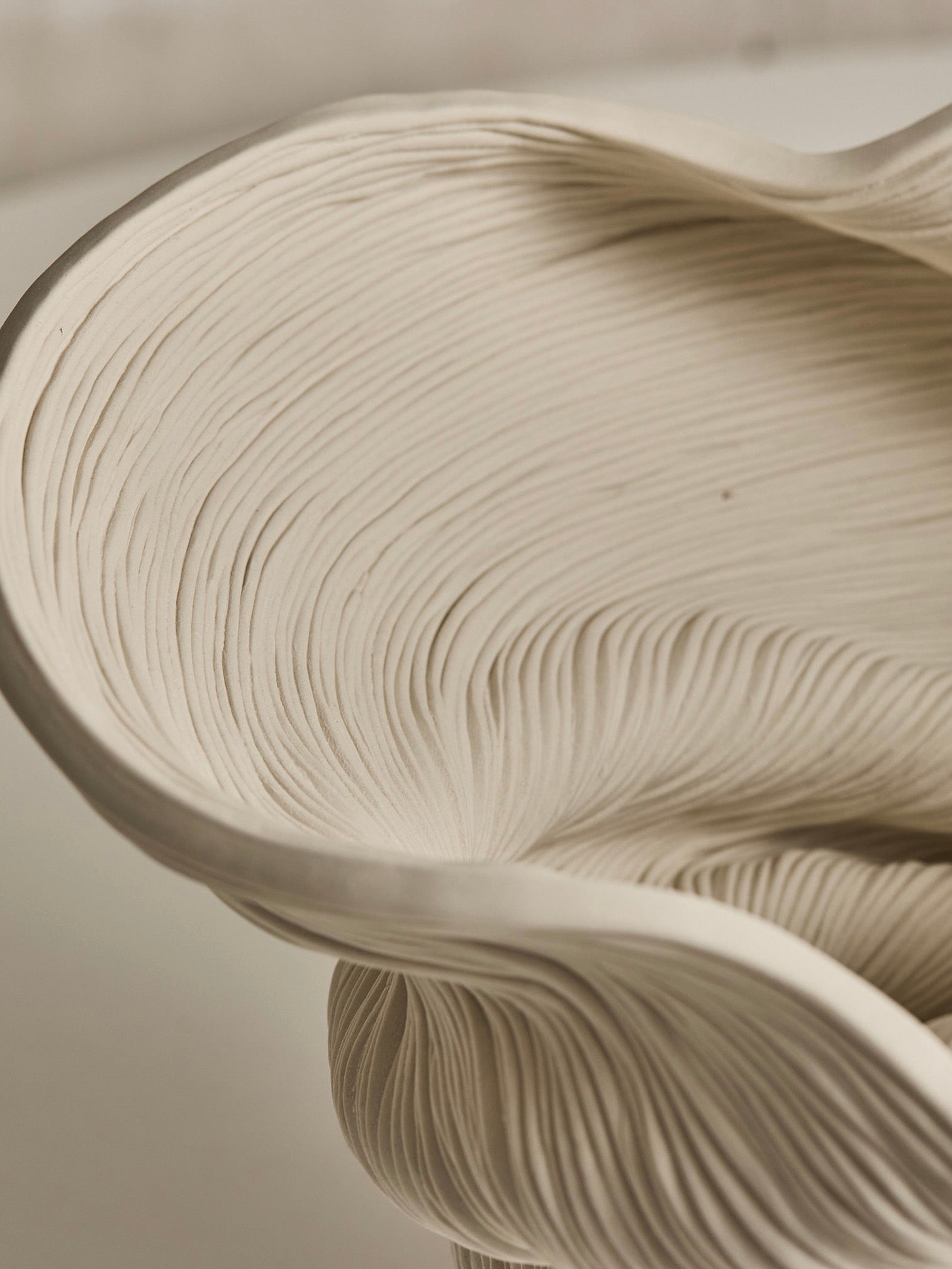 Close-up of 'Medium Flutter' by The Soucy Shop, a unique sculptural ceramic piece with intricate flowing grooves and organic textures. This handmade, smooth matte surface features curving, overlapping layers for depth and movement. Creamy beige porcelain coils add an elegant touch.