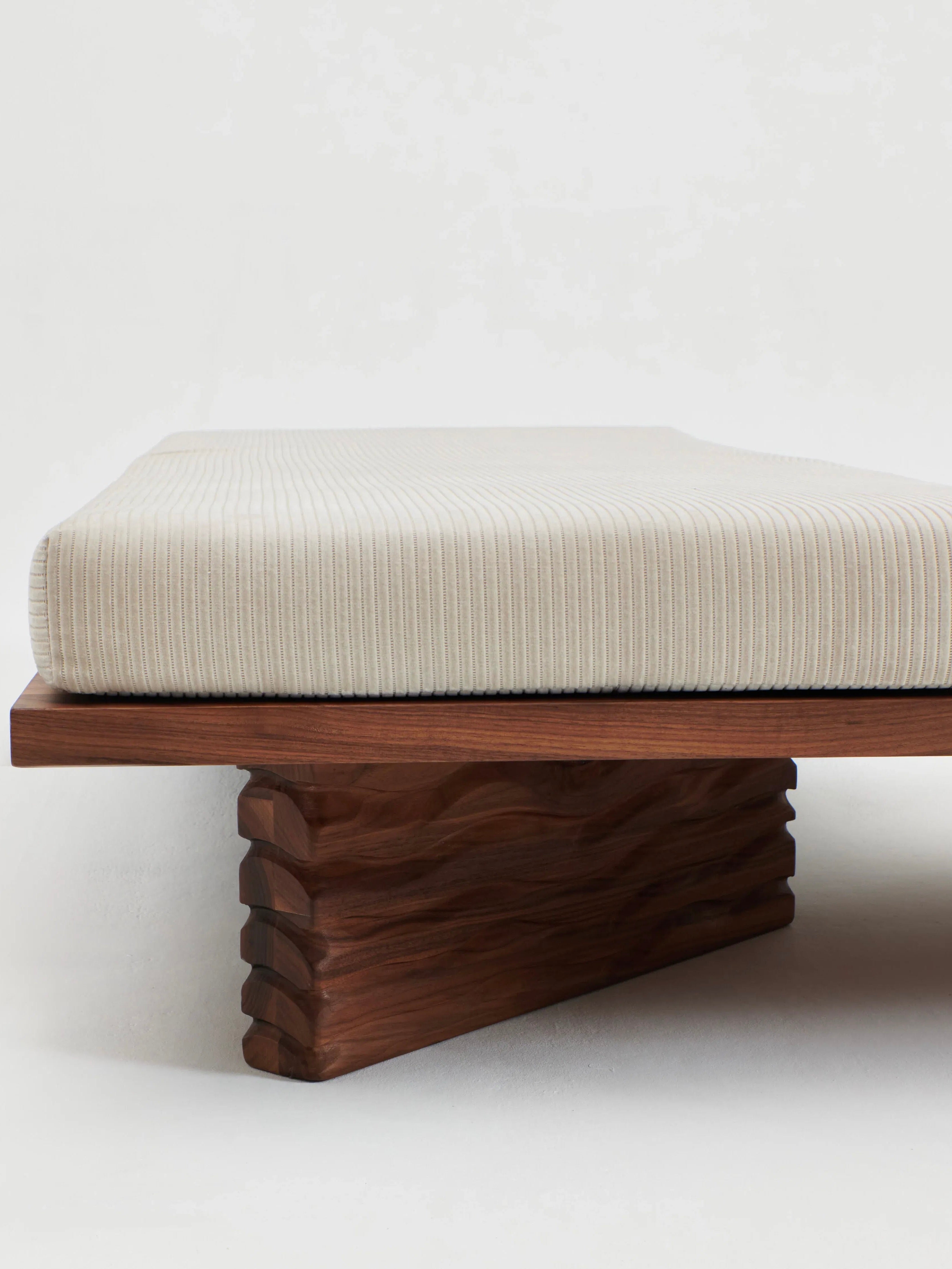 The Nazaré Daybed L Shape by Project 213A is a minimalist wooden bench featuring a textured, triangular base that supports a ribbed, light beige cushion. Handmade in Portugal from solid walnut, this design emphasizes clean lines and the natural grain of the wood, blending modern aesthetics with traditional craftsmanship.