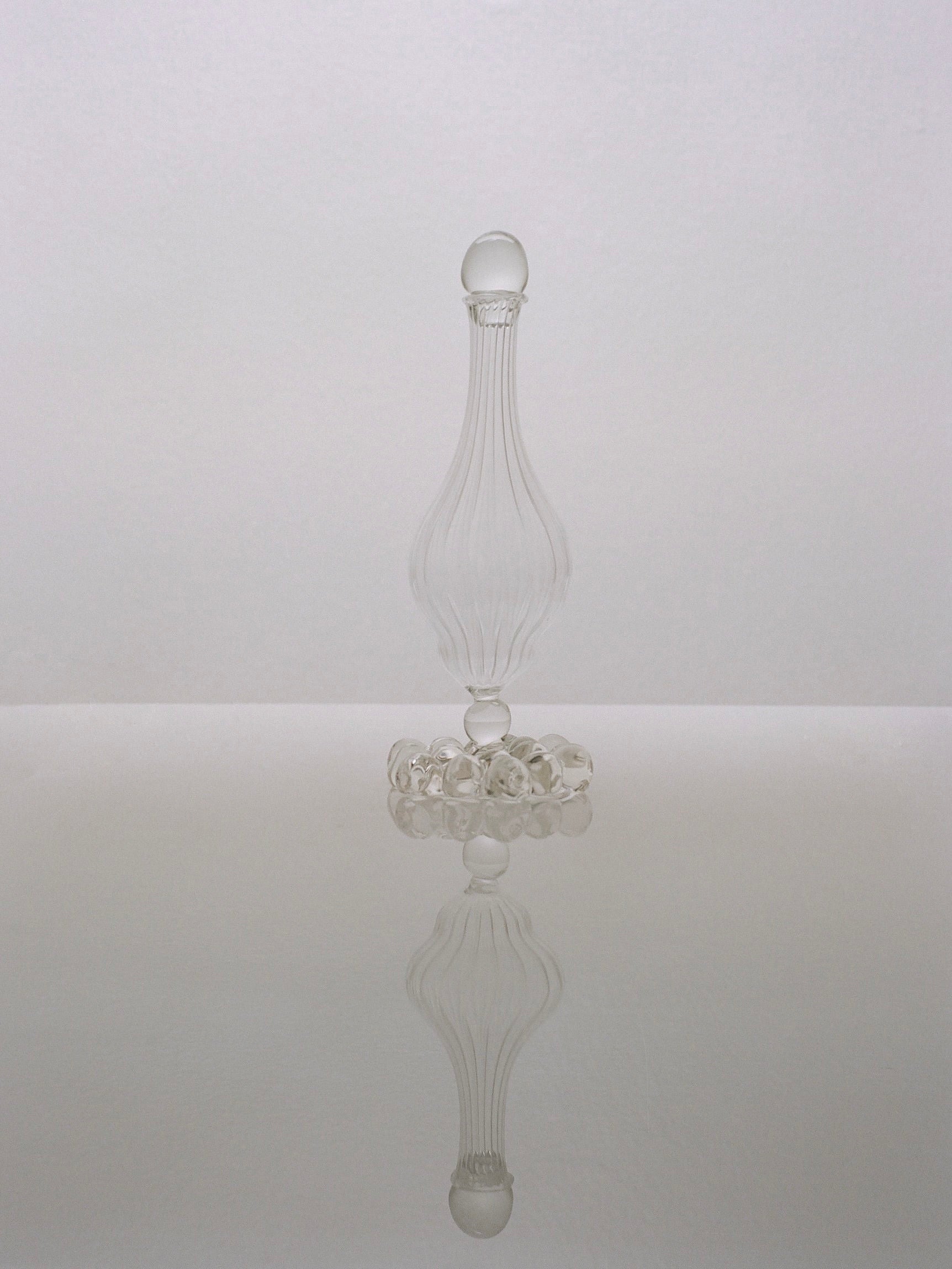 A clear, intricately designed glass bottle with a spherical stopper stands against a plain light backdrop. This artisanal piece, known as the Calypso Carafe by Justine Menard, boasts a slender, elegant form and is reflected perfectly on the surface beneath it.