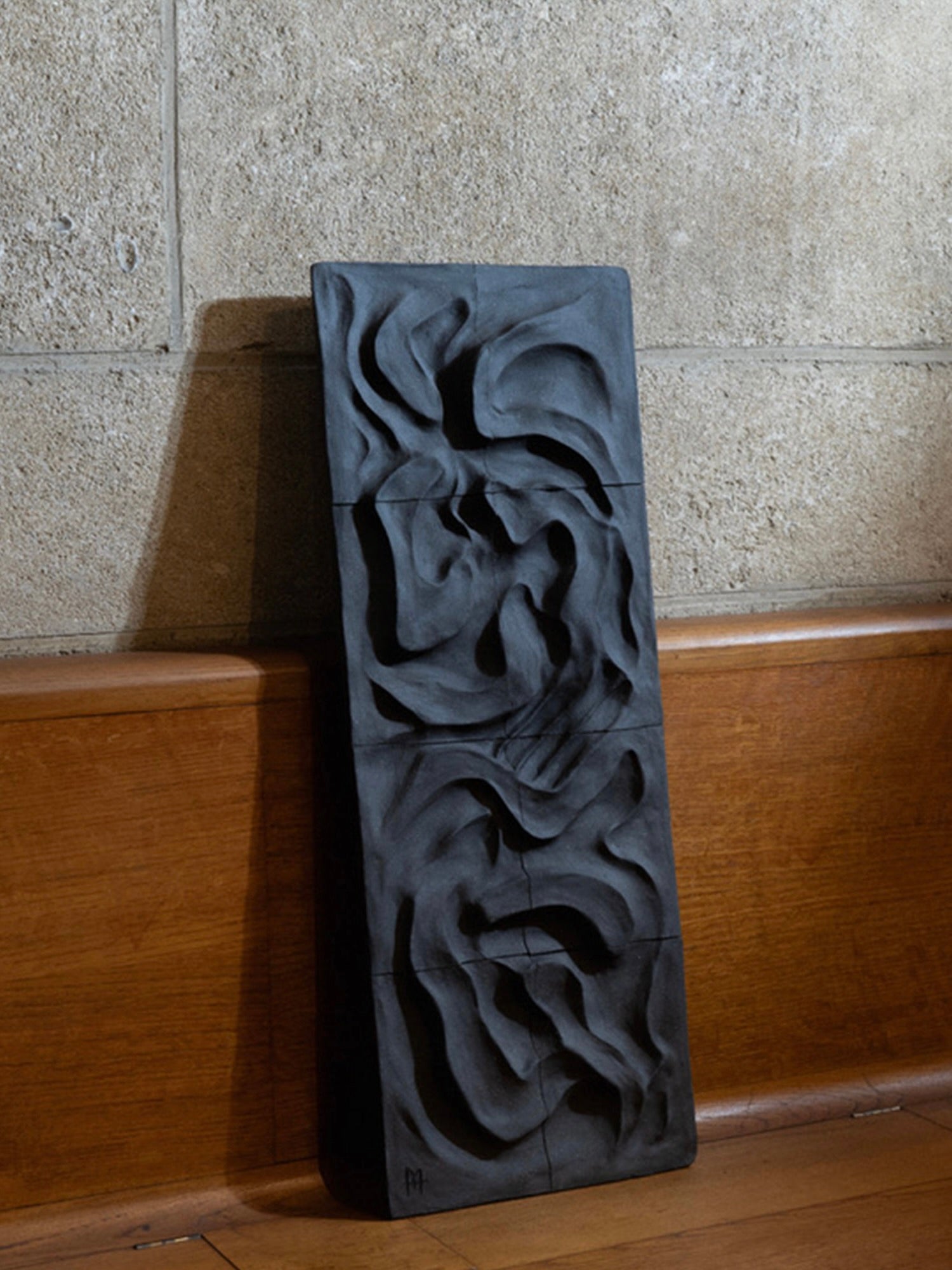 The Kilauea Lava wall sculpture by Franck Scala features black abstract patterns against a stone-textured wall and wooden surface, evoking ceramic bas-reliefs.