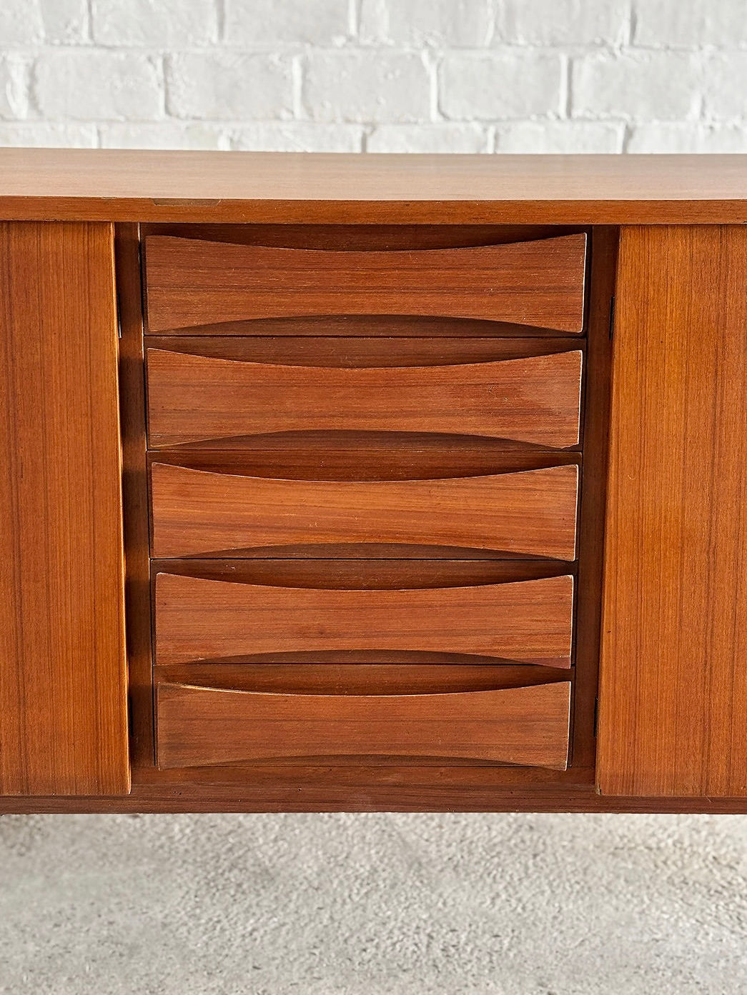 The Introverso Italian Modernist Teak Sideboard, designed by Vittorio Dassi in the 1960s, features five curved pull drawers and two vertical cabinet doors. Its warm teak finish contrasts beautifully against a white brick wall.