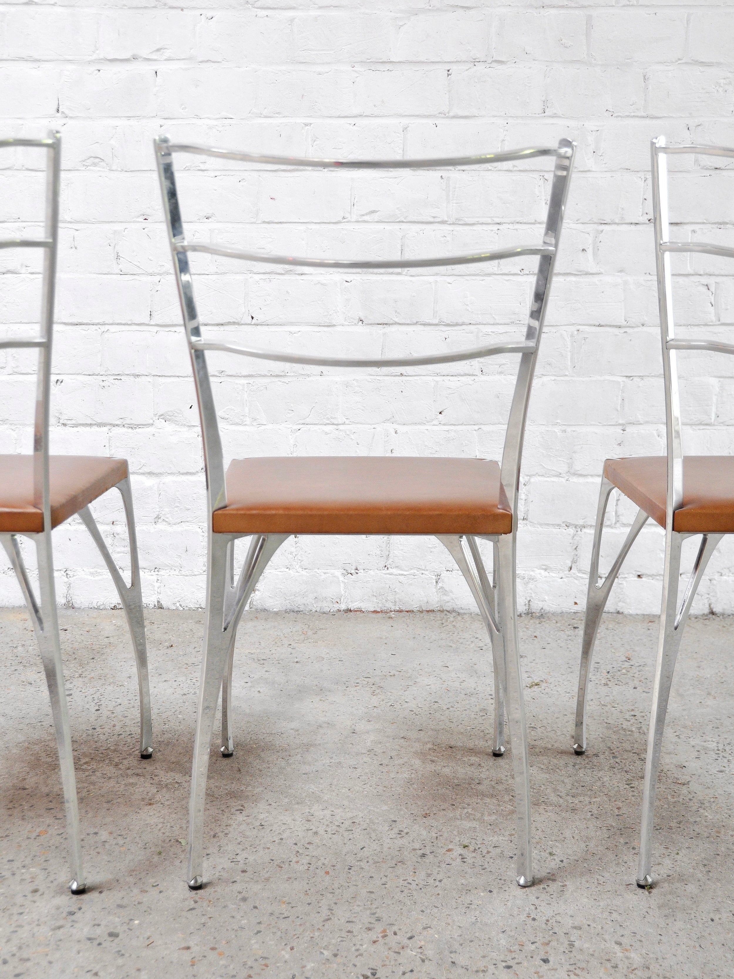 A row of three 'Organic' dining chairs by Erwan Boulloud from Introverso, featuring sculptural aluminum frames and brown seats, are arranged on a concrete floor against a stark white brick wall.