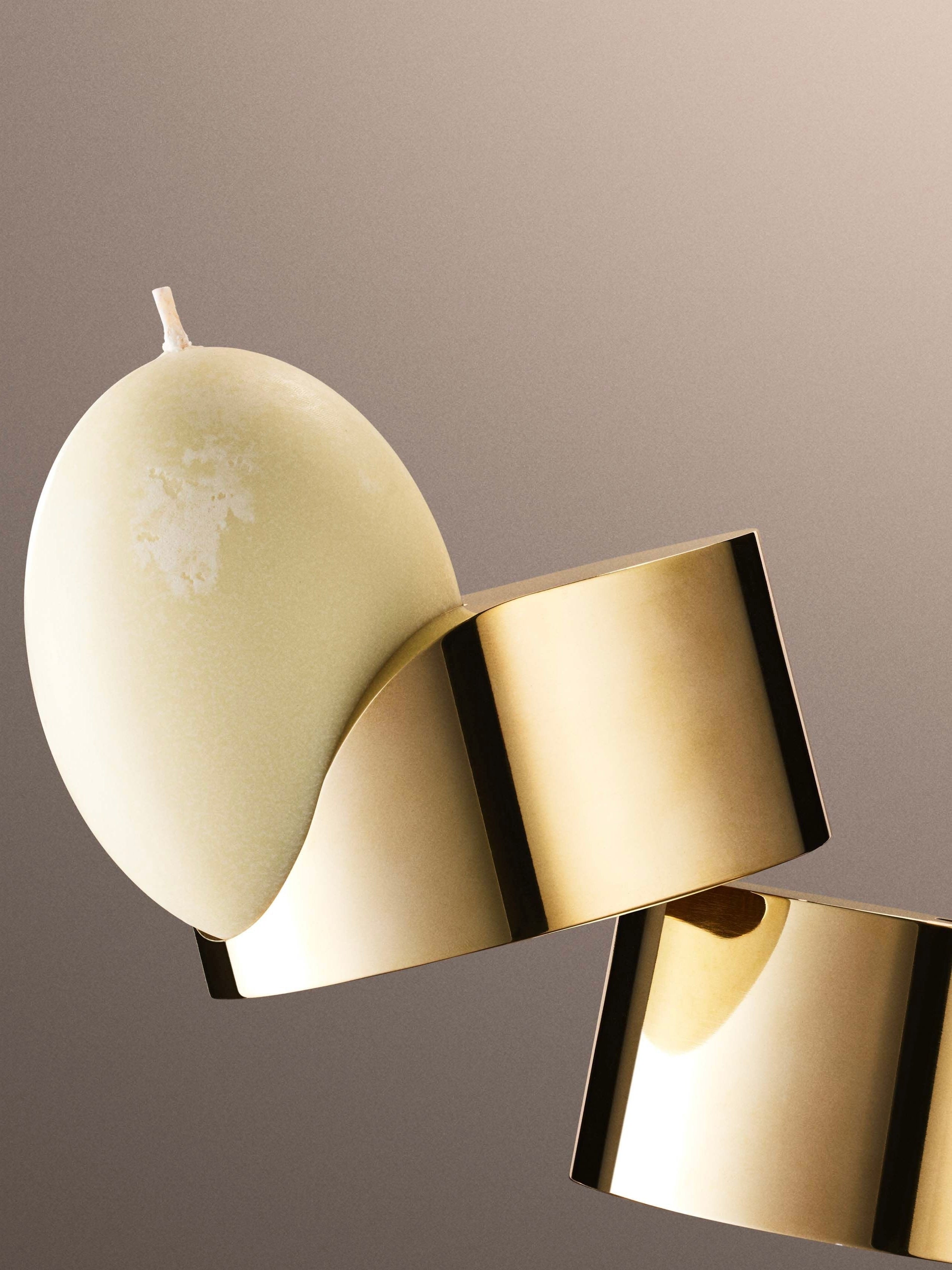 A cream-colored, egg-shaped candle alight on a Tête Studio Low Perch Candle Holder, set against a blurred beige background. The candle's off-center placement evokes the delicate balance of a Noguchi sculpture, nestled perfectly on the shiny gold metallic surface.