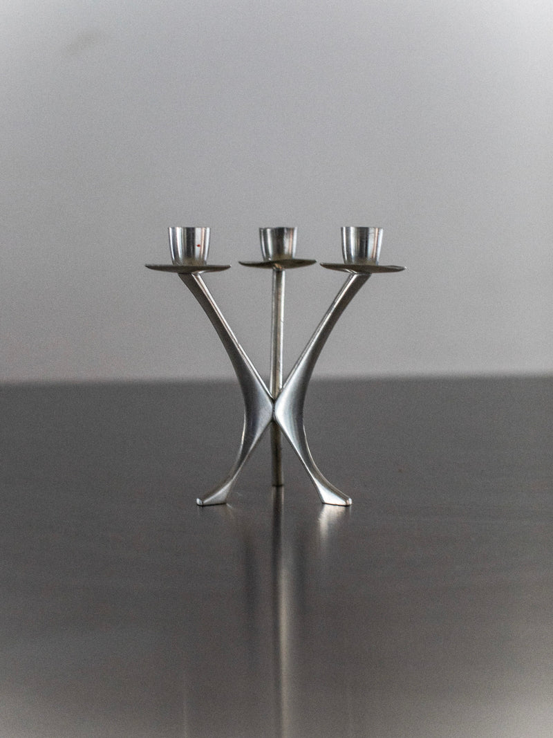 The Out For Lunch Steel Candleholder boasts a triangular design with three branches, resting on a reflective surface. Its sleek curves and symmetrical form embody contemporary decor, offering a stylish, modern aesthetic.