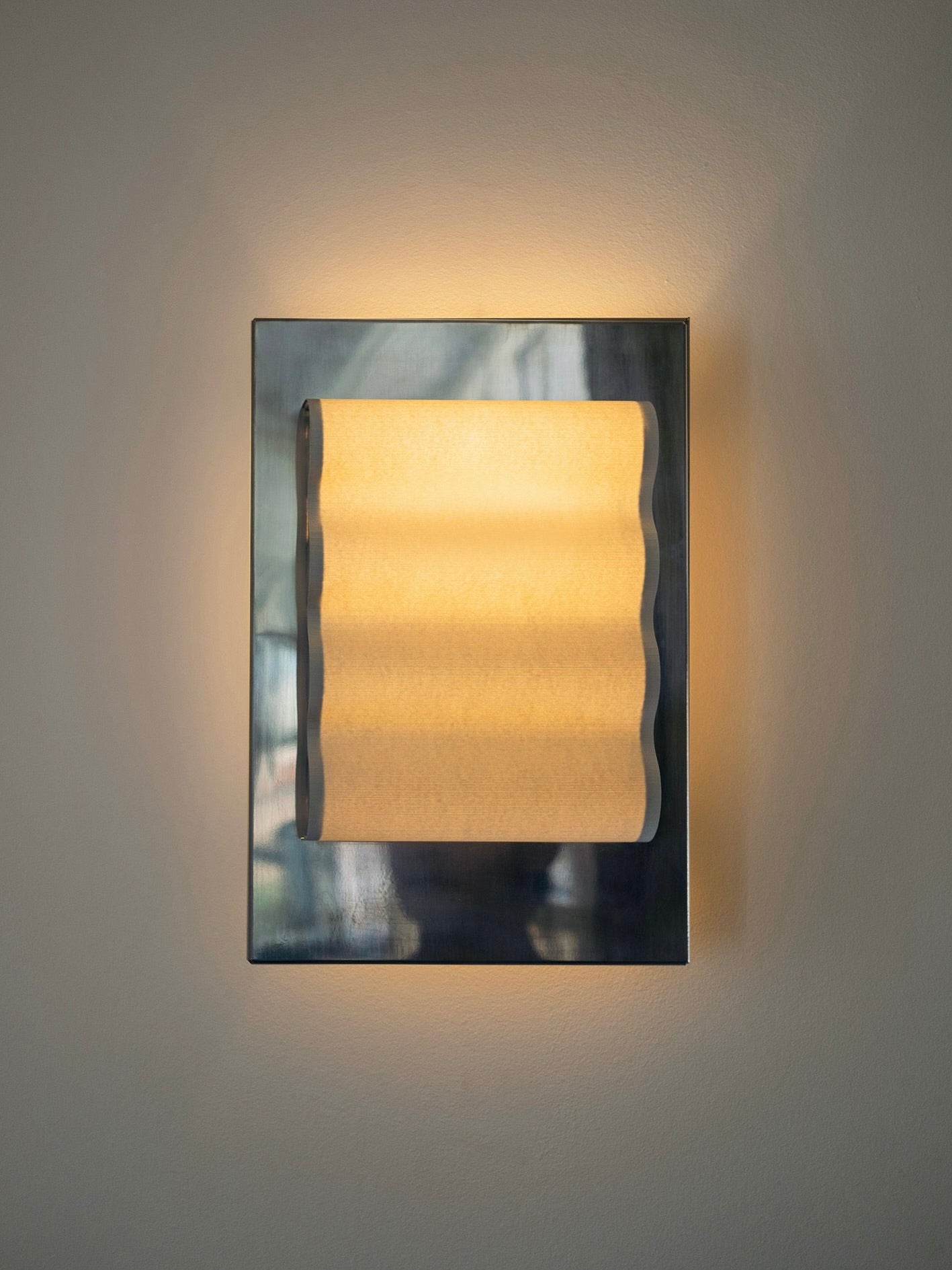 The Wall Light Frame Miroir M by Violaine d'Harcourt is mounted on a light beige wall. This modern wall sconce features a metallic frame and a diffused wavy light shade, embodying minimalist design. The handcrafted wall sconce emits a warm light, creating a soft, ambient glow around it.