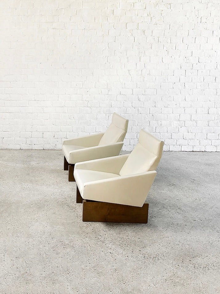 Two Introverso French Modernist lounge chairs, crafted in white eco-leather upholstery with wooden bases, are placed side by side on a light gray concrete floor. Nestled against a white brick wall, they evoke a clean and minimalist aesthetic of the 1950s.