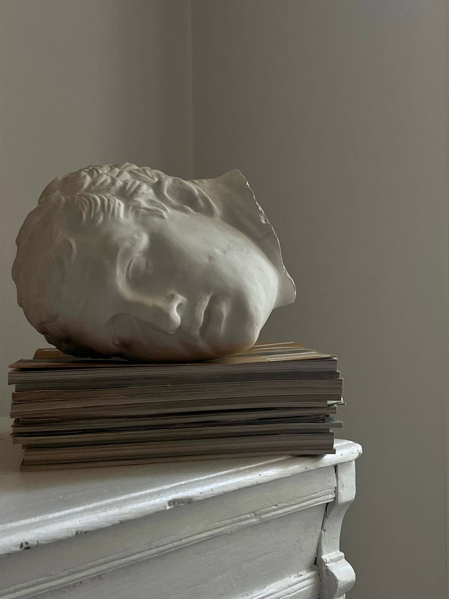 The Italian Male in Plaster by Médecine lies on its side atop a stack of magazines or books on a white wooden surface. The scene is dimly lit with muted tones, creating a calm and minimalist atmosphere that accentuates the vintage design of the durable plaster sculpture.