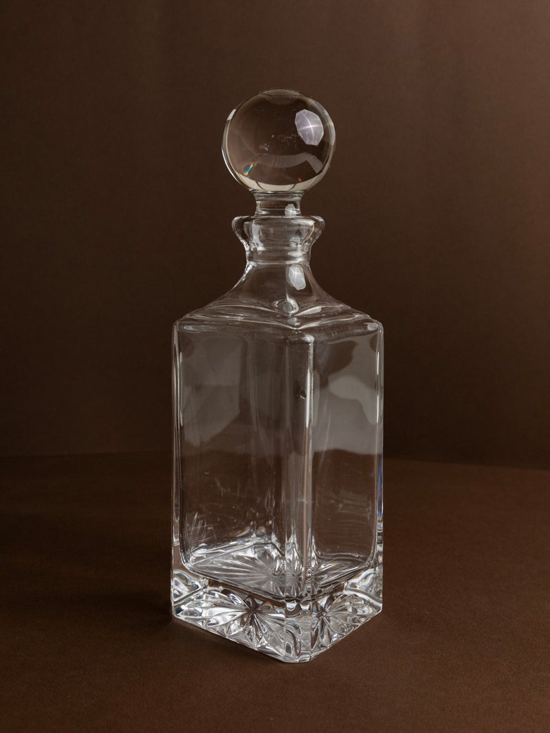 The Murano Glass Carafe 70s by Bottega Jacobs, featuring a clear vintage design with a round stopper, is set against a dark brown backdrop. The carafe's reflective surface beautifully captures the light, highlighting its geometric shape and sophisticated design.