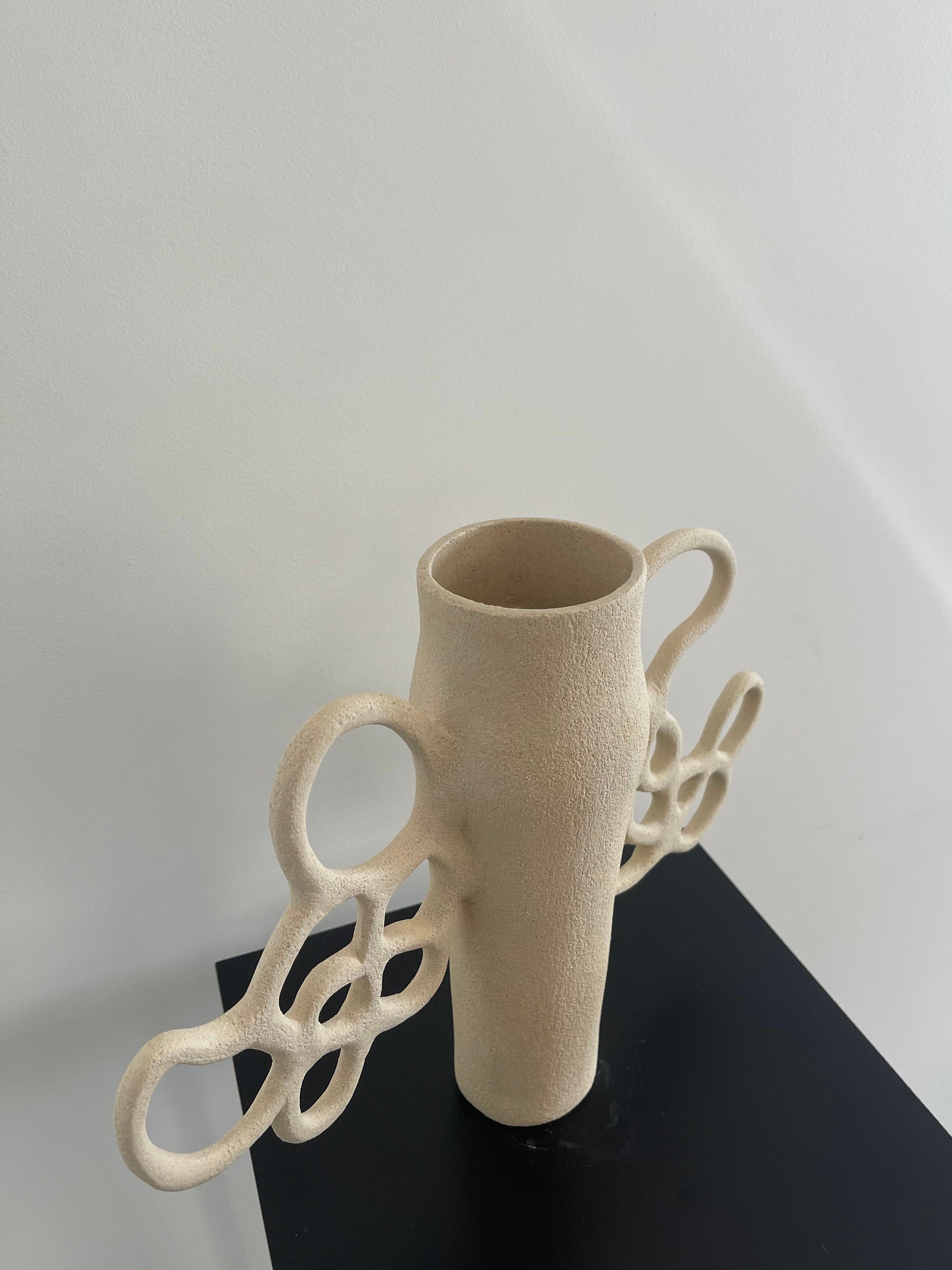 As a member of the Liens collection by Louana Roudot Ceramic, the Vase Liens n°1 in beige ceramic is crafted from white chamotte stoneware, featuring an elongated body with intricate looped handles that resemble abstract wings. It is elegantly displayed on a dark surface against a light background.