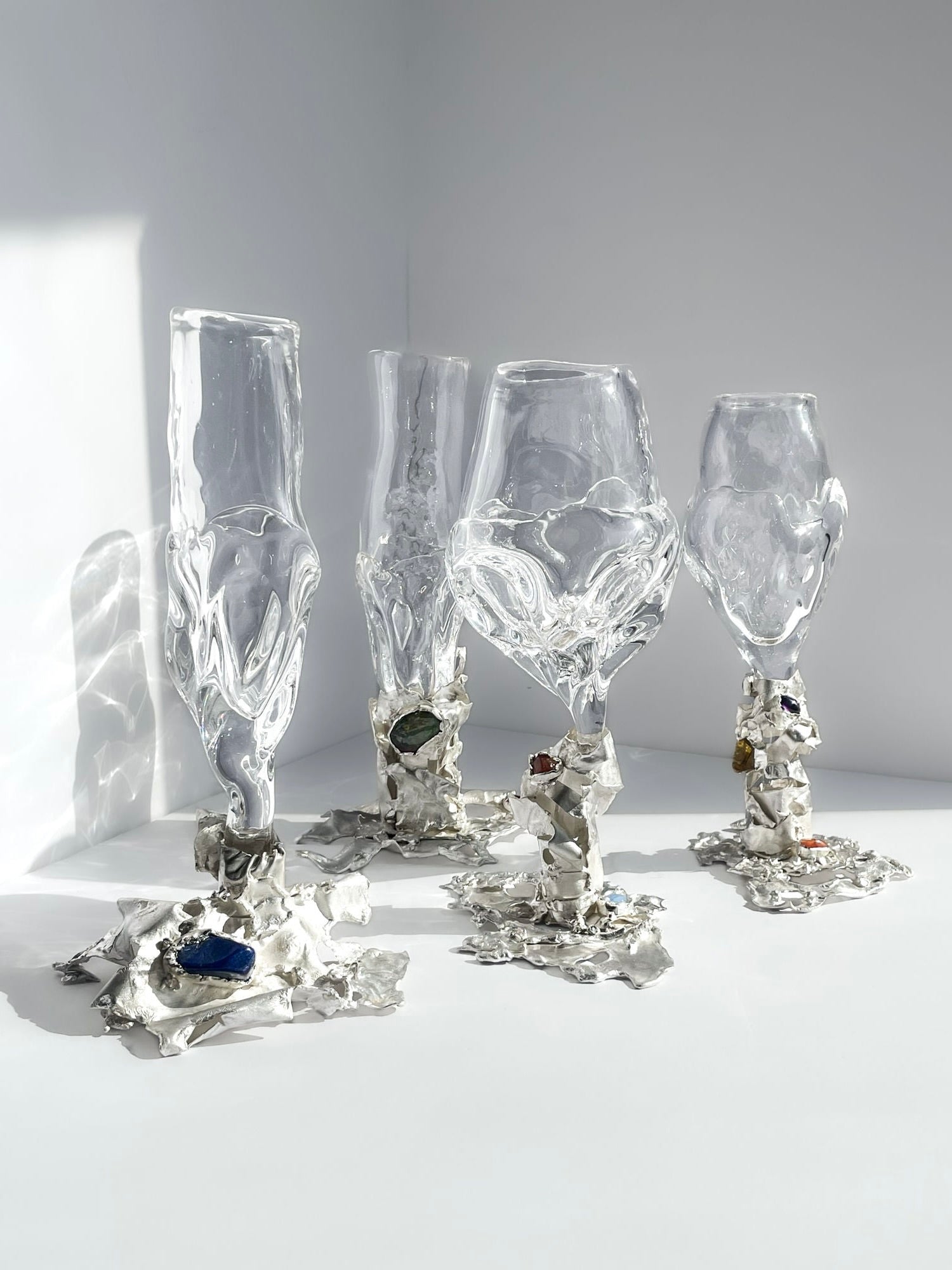 Four artistic Jewelled Goblet no. 10s by Lindsey Fontijn from Szkło Studio stand on a flat surface, each featuring uniquely distorted shapes with intricate, irregular bases adorned with colorful natural gemstones. The goblets are arranged under soft lighting, casting shadows on a plain, light gray background.