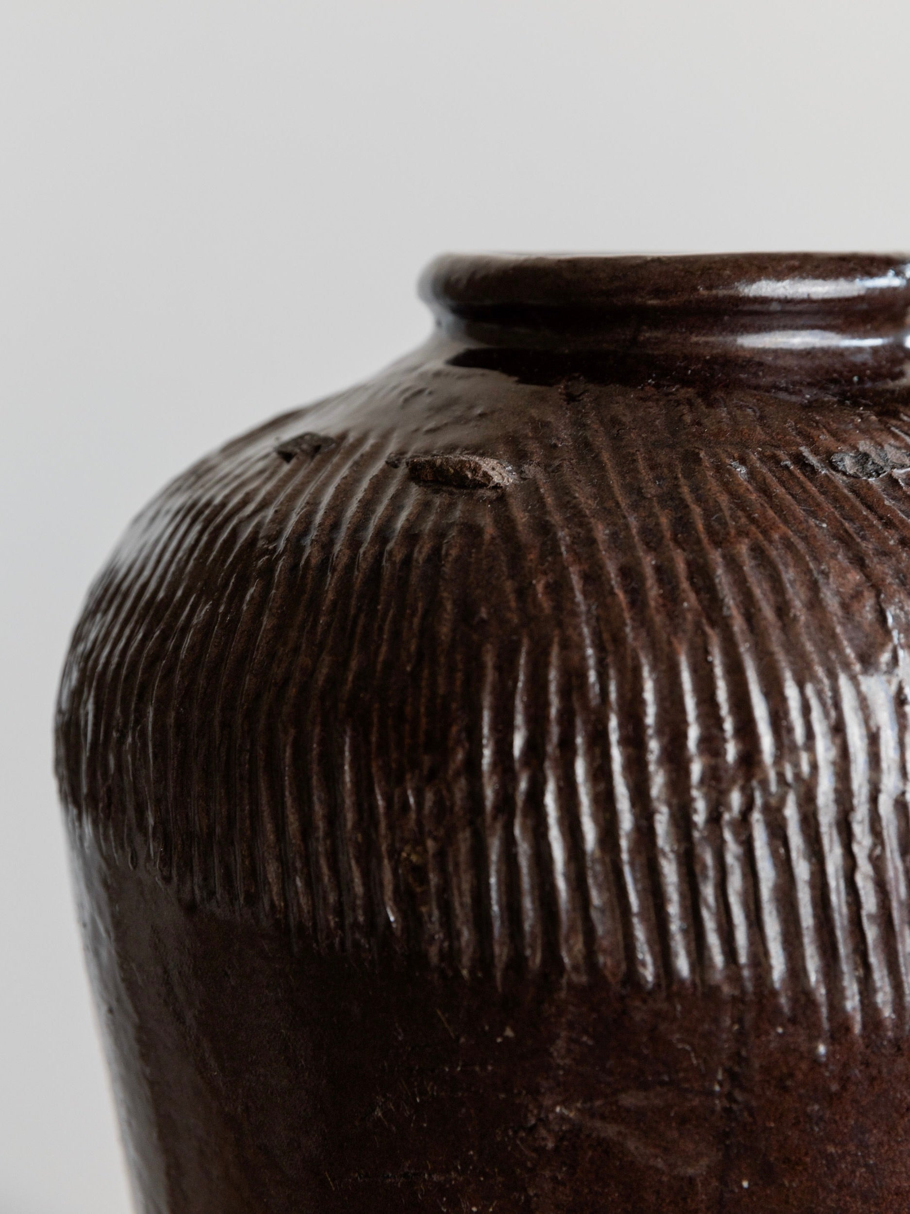 Close-up of the Bottega Jacobs Antique Chinese 19th Century Ceramic vase featuring a dark brown shade with a glossy finish. Its slightly flared neck and vertically grooved textured pattern add depth to its smooth surface, evoking the look of an ancient Chinese rice wine pot. The background is softly blurred.