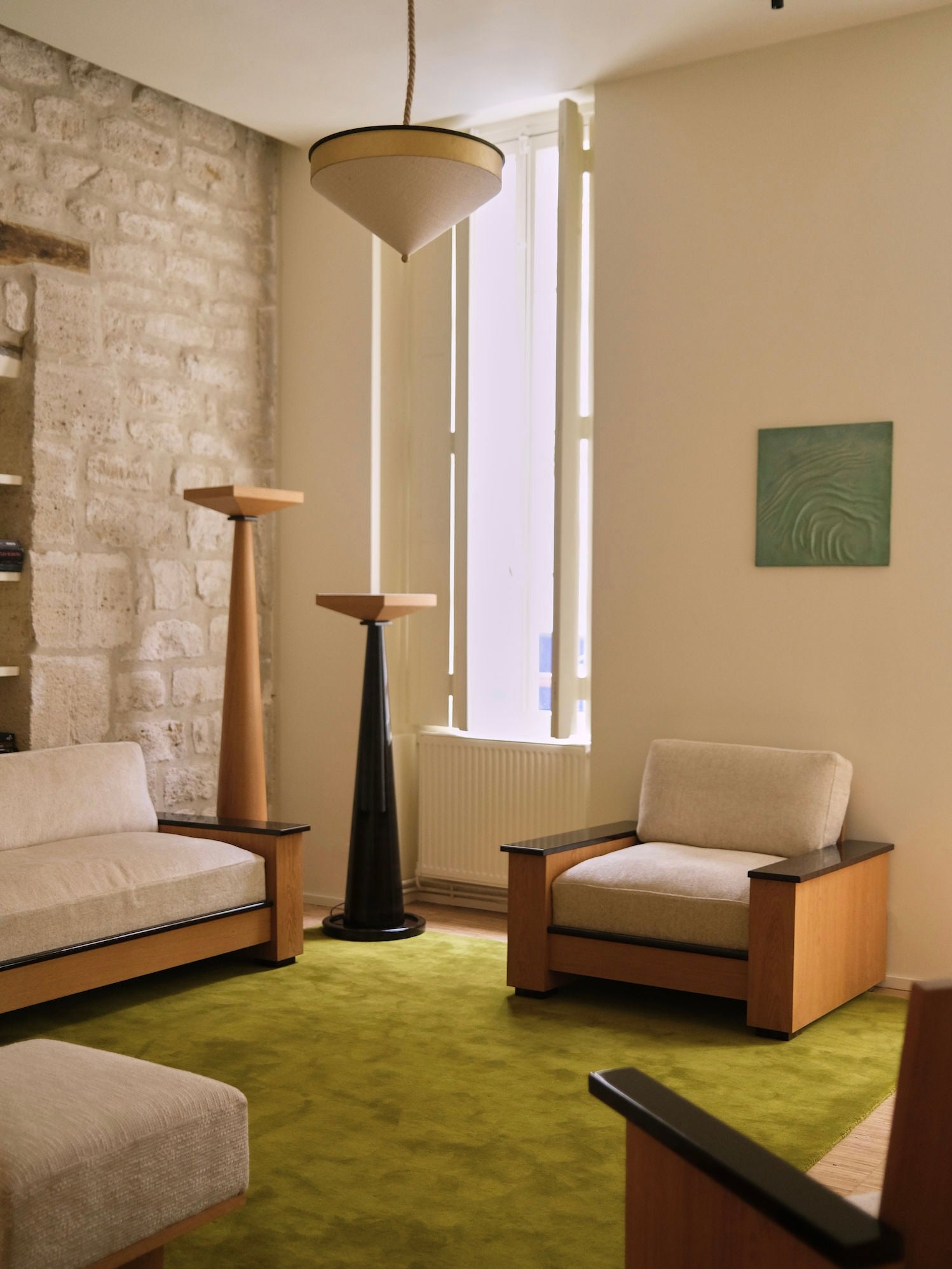 A cozy living room is adorned with two modern armchairs resting on a green carpet. The "Le Fontainebleau" Floor Lamp by HAUVETTE & MADANI, with its tall cone-shaped wooden design, beautifully complements the lacquered oak accents and pairs effectively with a unique ceiling light. A painting decorates the stone wall as natural light floods through the tall windows.