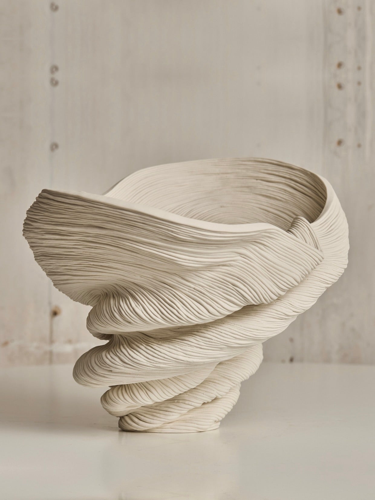 The Soucy Shop's Medium Flutter is a sculptural porcelain piece with swirling, twisted forms reminiscent of layered fabric. The off-white, textured design stands out against a neutral background, creating an impression of delicate dynamism.