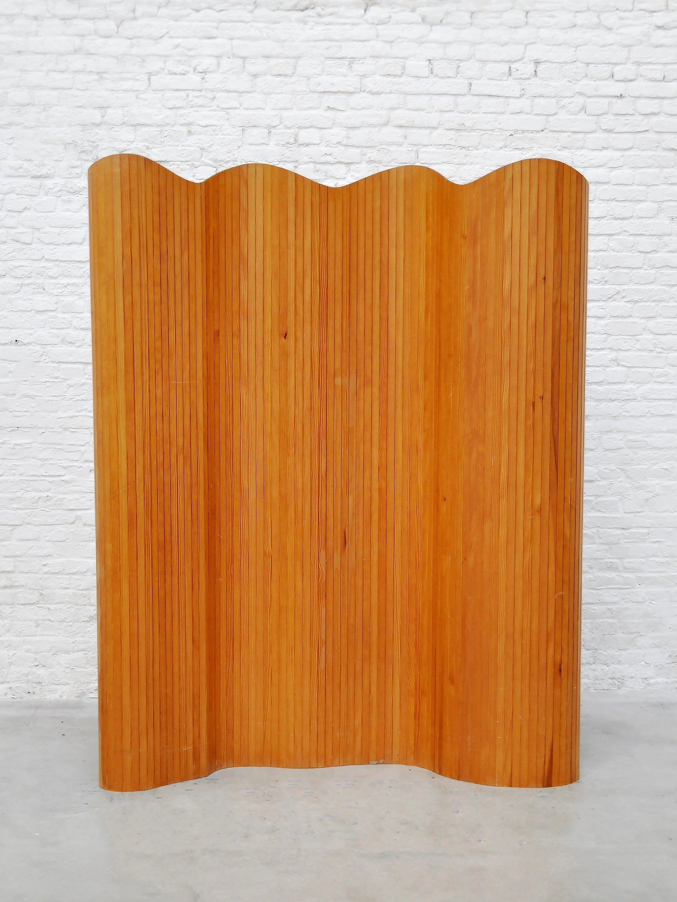 The Jomaine Baumann Room Divider by Introverso, crafted in the 1940s, stands against a white brick wall. Its French Art Deco elegance shines through with tambour-inspired grooves and gentle curves for an organic feel.