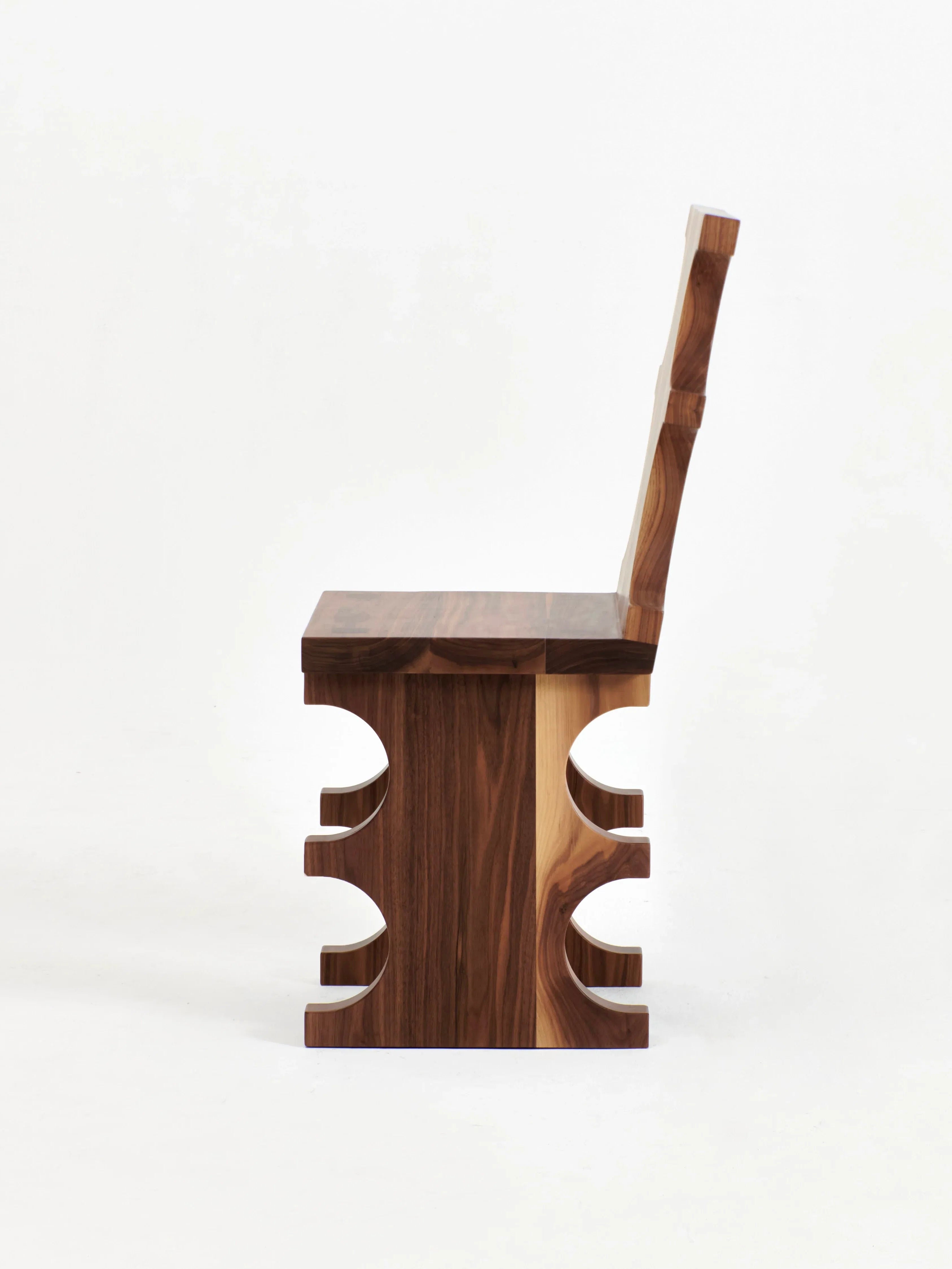 A side view of the Portugal Chair No 7 by Project 213A, crafted by skilled artisans from solid walnut, showcases its distinct geometric design. The chair features a solid backrest and seat, while the base has cut-out semicircle shapes on both sides, adding an artistic element. The entire piece is made from richly colored wood.