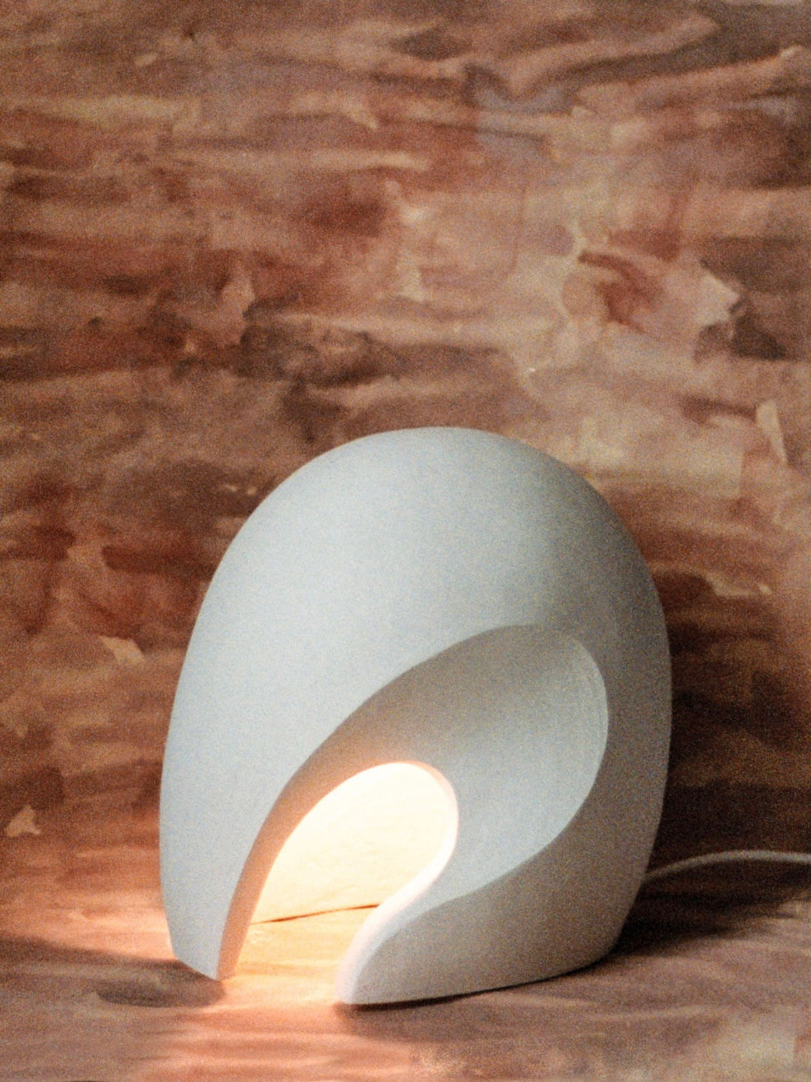 The Coquille - Ceramic Lamp by Lea Bigot, a small handmade white ceramic piece with a modern curved design, emits a warm glow against an earthy brown background, casting soft shadows.