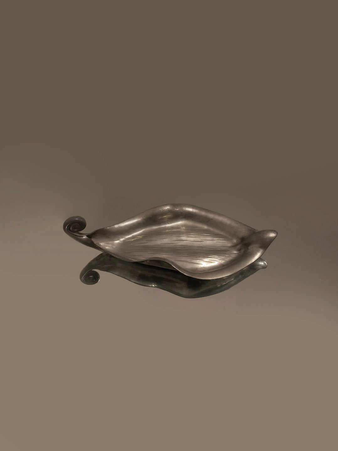 A Handmade Leaf Vide Poche by Les Objoies rests on a reflective surface against a plain gray backdrop. Its pewter finish gleams, highlighting its elegant contours and smooth texture, with curled handle edges capturing the essence of an Art Nouveau design.