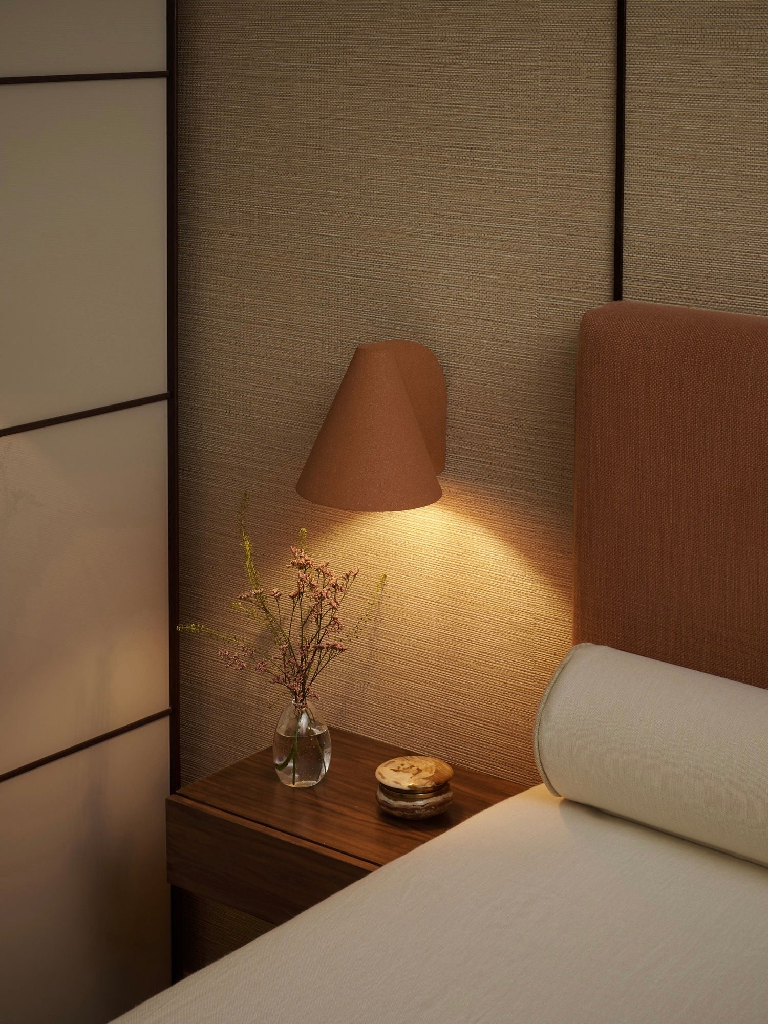 A cozy bedroom corner boasts a brown upholstered bed and a wooden bedside table with a handcrafted stoneware vase. The 'Esoteric' Almond Brown Wall Light from Mariza Galani casts soft, warm light on the textured wall, creating an inviting ambiance.