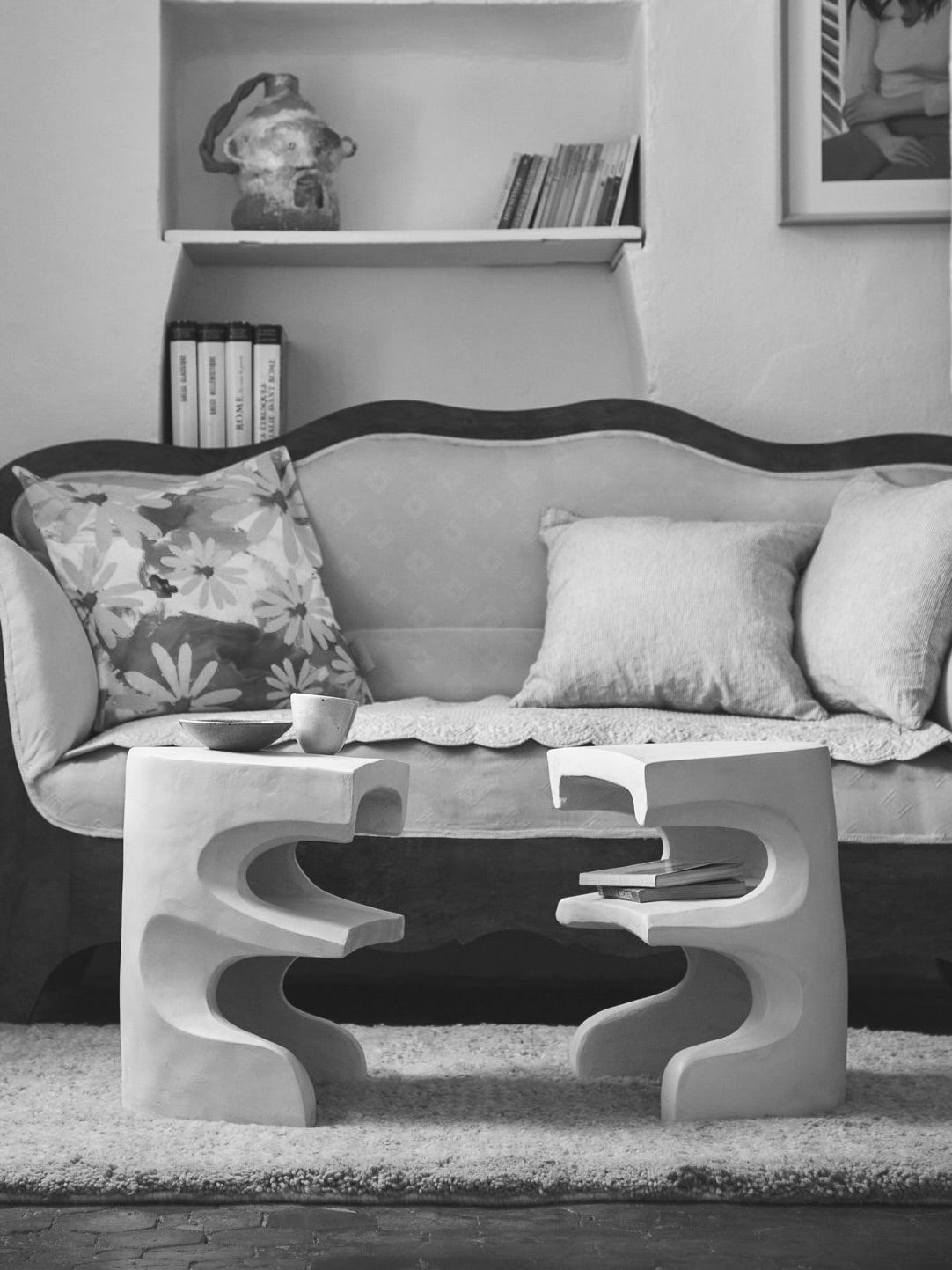 A black and white image of a cozy living room features a vintage sofa with pillows and the Baisers Volés Carved Stoneware Coffee Table by Lea Bigot. The uniquely designed table, with abstract curves and shelves, rests on a textured rug displaying handmade cups and decor items.