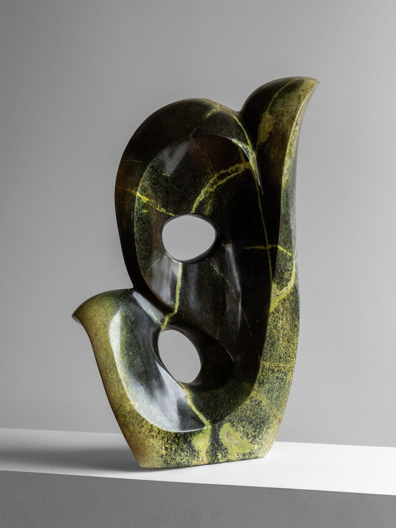 Abstract Stone Sculpture II by Spigel is a dark green sculpture with smooth curves and circular openings, crafted from leopard stone with lighter green veins. It boasts a glossy finish and rests on a white surface against a gray background.