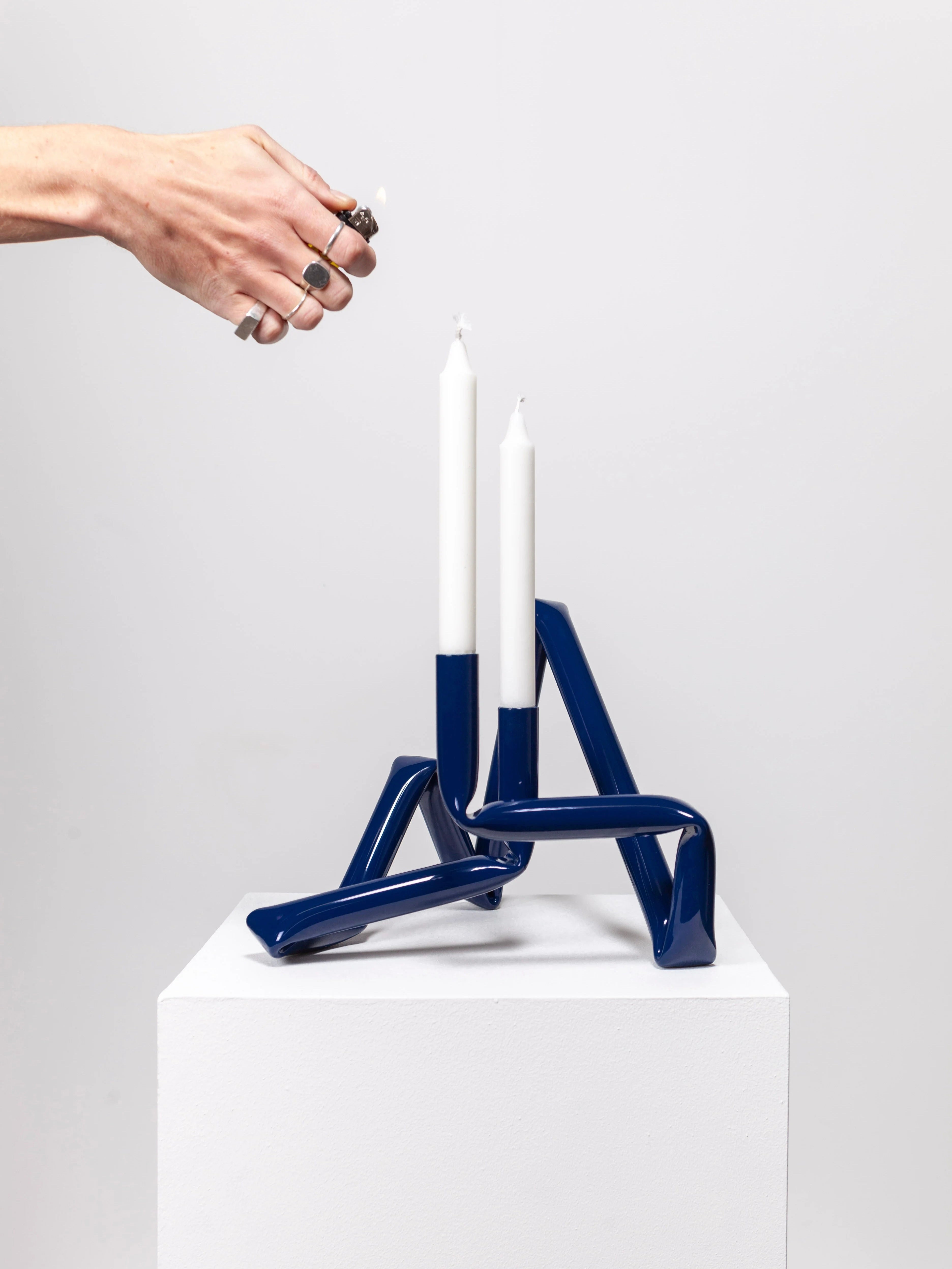 A hand adorned with rings holds a lighter near two white candles, which are mounted on an abstract, midnight blue Bucatini Candle Holder from AOT STUDIO. This sophisticated piece, handcrafted in Denmark from sleek aluminum tubing, rests elegantly on a white pedestal against a plain white background.