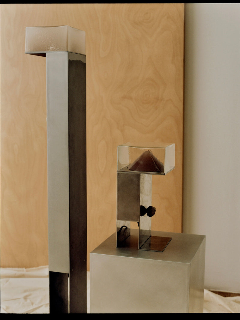 Two modern sculptures decorate a neutral background. The taller, Perceptions Floor Lamp by Known Work, resembles an atmosphere with a narrow metallic column and transparent top. The shorter piece has a metallic square base supporting a hand-blown glass container containing a small pyramid.