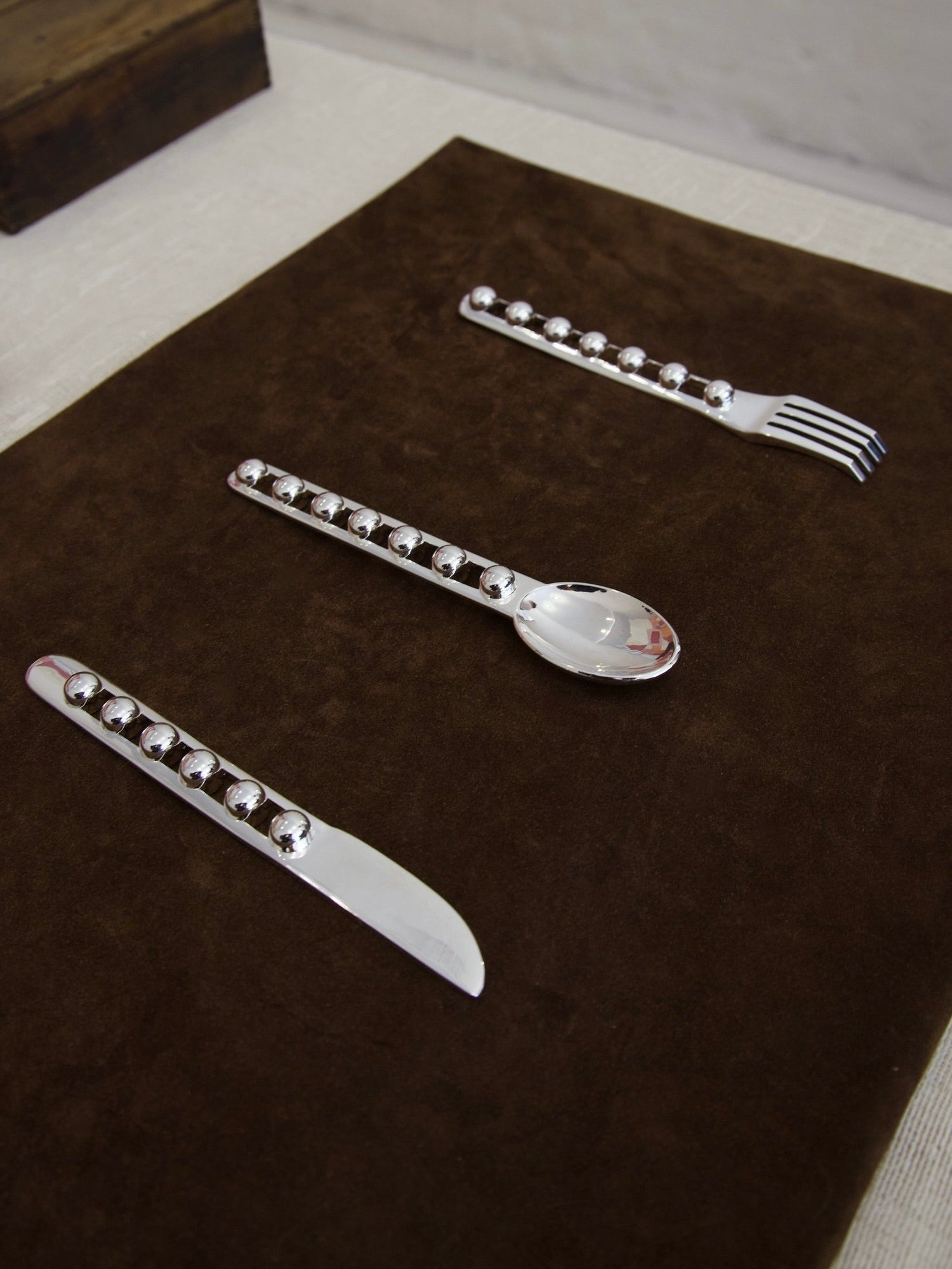 The "Seven Ball" Cutlery Set by Jessi Burch is elegantly displayed on a brown placemat. This functional art piece includes a knife, spoon, and fork, each adorned with decorative round accents along the handles.