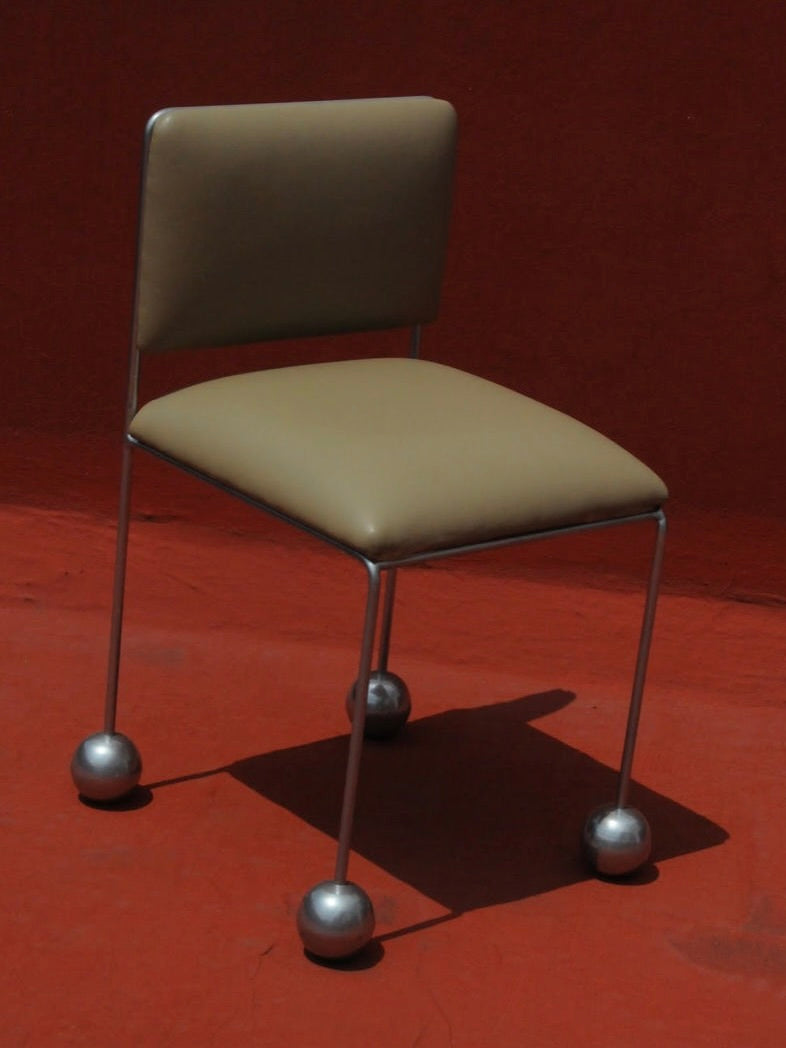 Introducing the Panorammma 'Ball Foot' Chair, showcasing a light brown vegan textile cushioned seat and backrest, complemented by a minimalist metal frame with distinctive spherical feet set against a vivid reddish background.