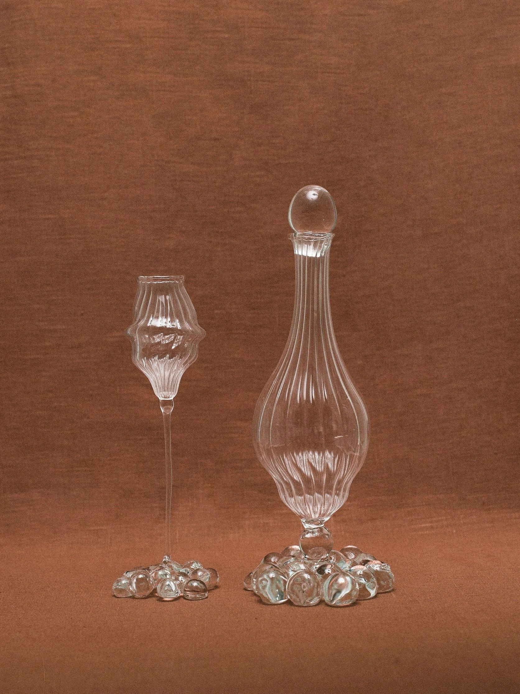 A clear glass goblet with a textured design stands next to the elegant Calypso Carafe by Justine Menard, an artisanal piece featuring a rounded stopper. Both are placed on a brown fabric background and encircled by scattered clear glass marbles, showcasing the beauty of hand-blown glass artistry.