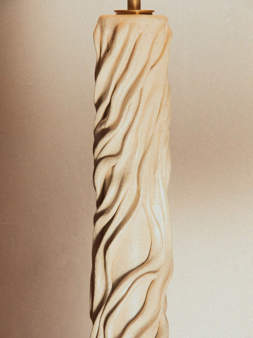 The "Waves" Table Lamp by Lorea Palacios features a tall, intricately carved cream-colored design with organic patterns and a wavy, textured surface that complements contemporary interiors. Its elegant, sculptural appearance resembles a ceramic masterpiece with a golden holder at the top.