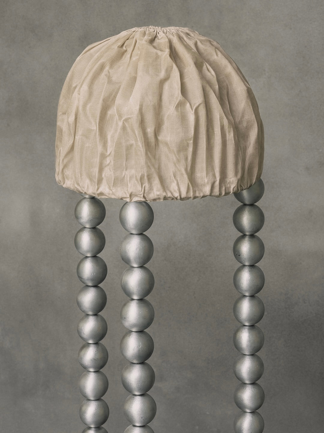 The "ALTER" Floor Light by Kaytar is a handcrafted, jellyfish-inspired lamp with a beige, crumpled linen dome supported by four legs made of sand-cast aluminium spheres set against a mottled gray beeswax backdrop.