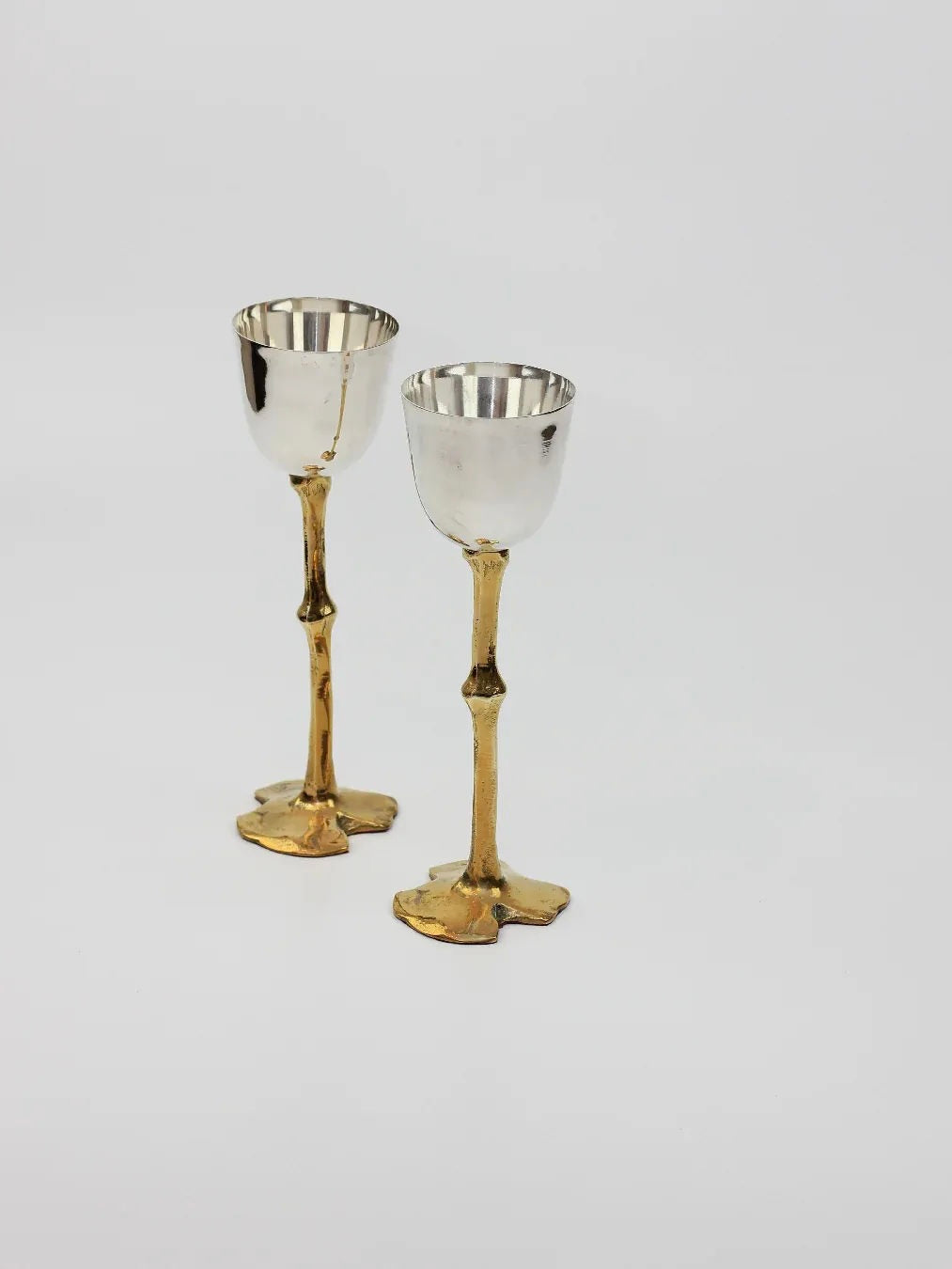 Two elegant goblets from Boga Avante Shop, featuring shiny silver cups and brass stems, stand on a light background. Hand-sculpted with a unique textured design, these Brutalist Goblets exude a sophisticated appearance.