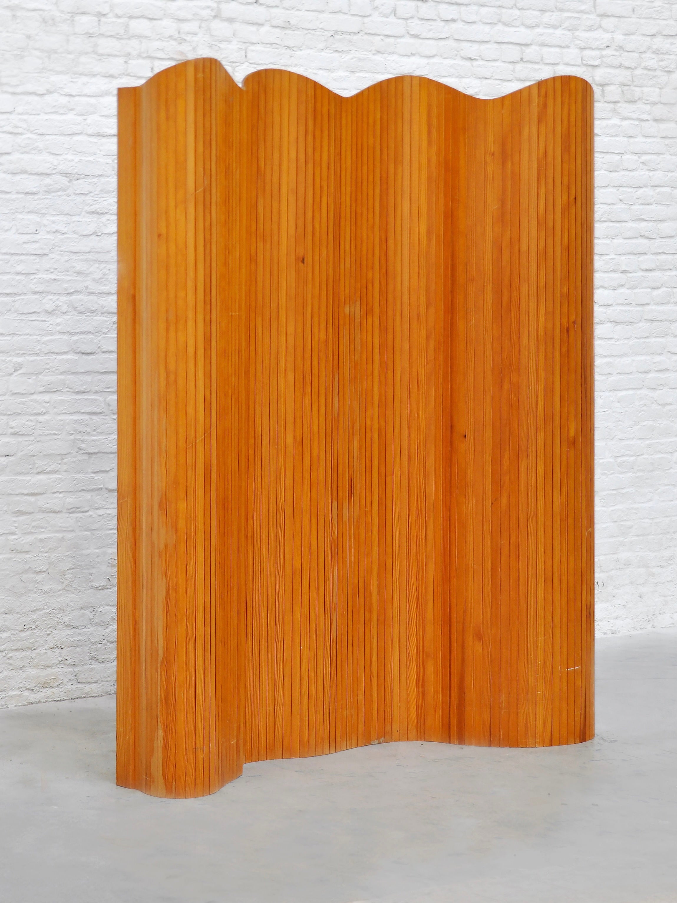 The Jomaine Baumann Room Divider by Introverso stands against a white brick wall, featuring 1940s French Art Deco style with wavy tambour vertical slats and a natural wood finish for a minimalist yet elegant look.