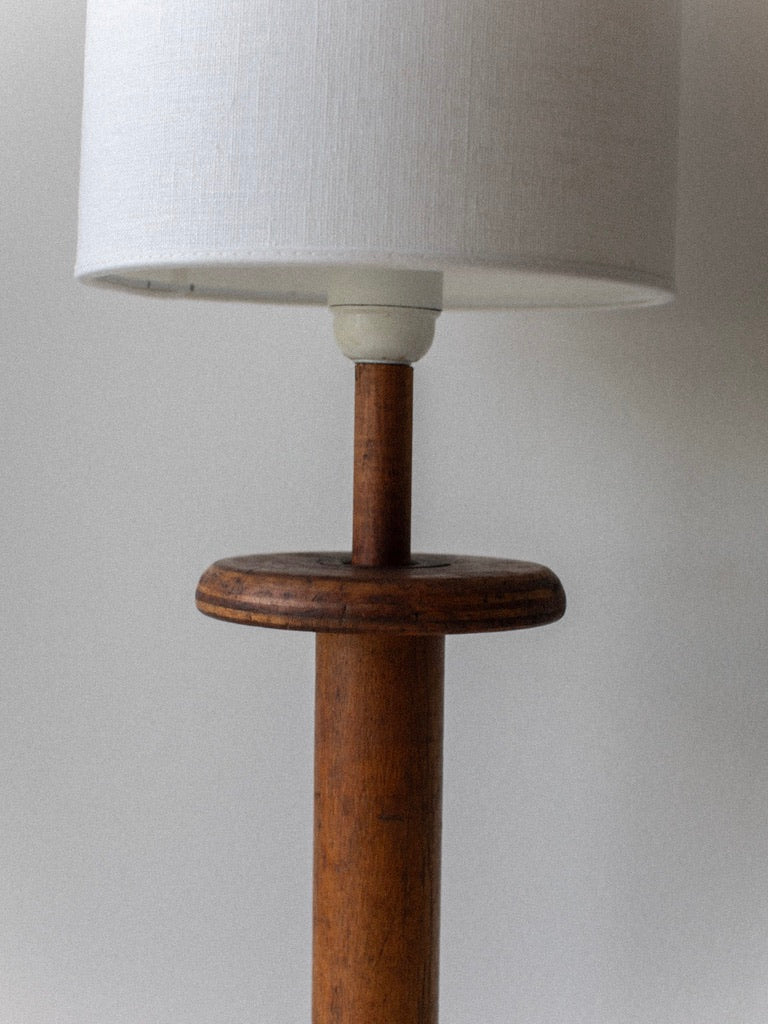 Introducing the Out For Lunch Midcentury Table Lamp, a rare find featuring a wooden handle base and a white fabric lampshade. The cylindrical base transitions seamlessly into a short wooden stem that supports the shade, resting on a slightly wider circular top against a plain, light-colored wall.