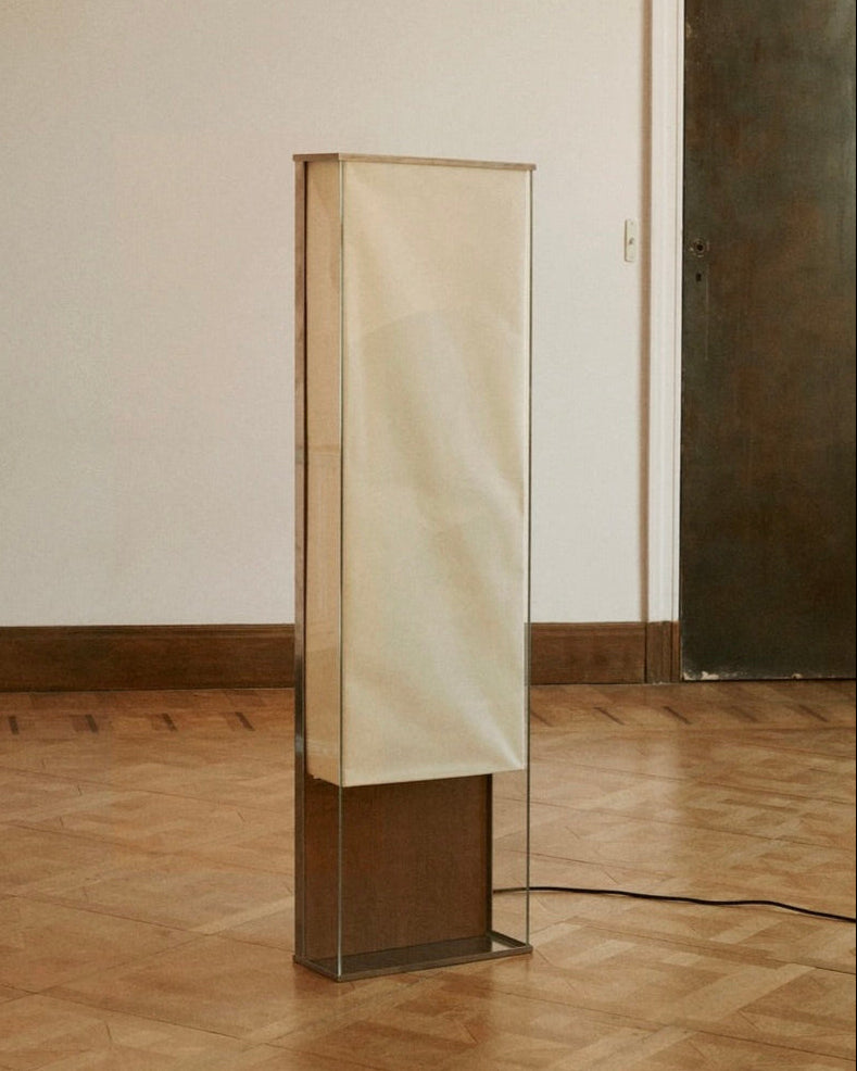 The Floor Lamp 2022 by Studio HAOS, featuring a tall rectangular minimalist design with a translucent cream shade and clear base, stands on a wooden parquet floor. A black power cord extends from its side; part of a sculpture appears on the left.
