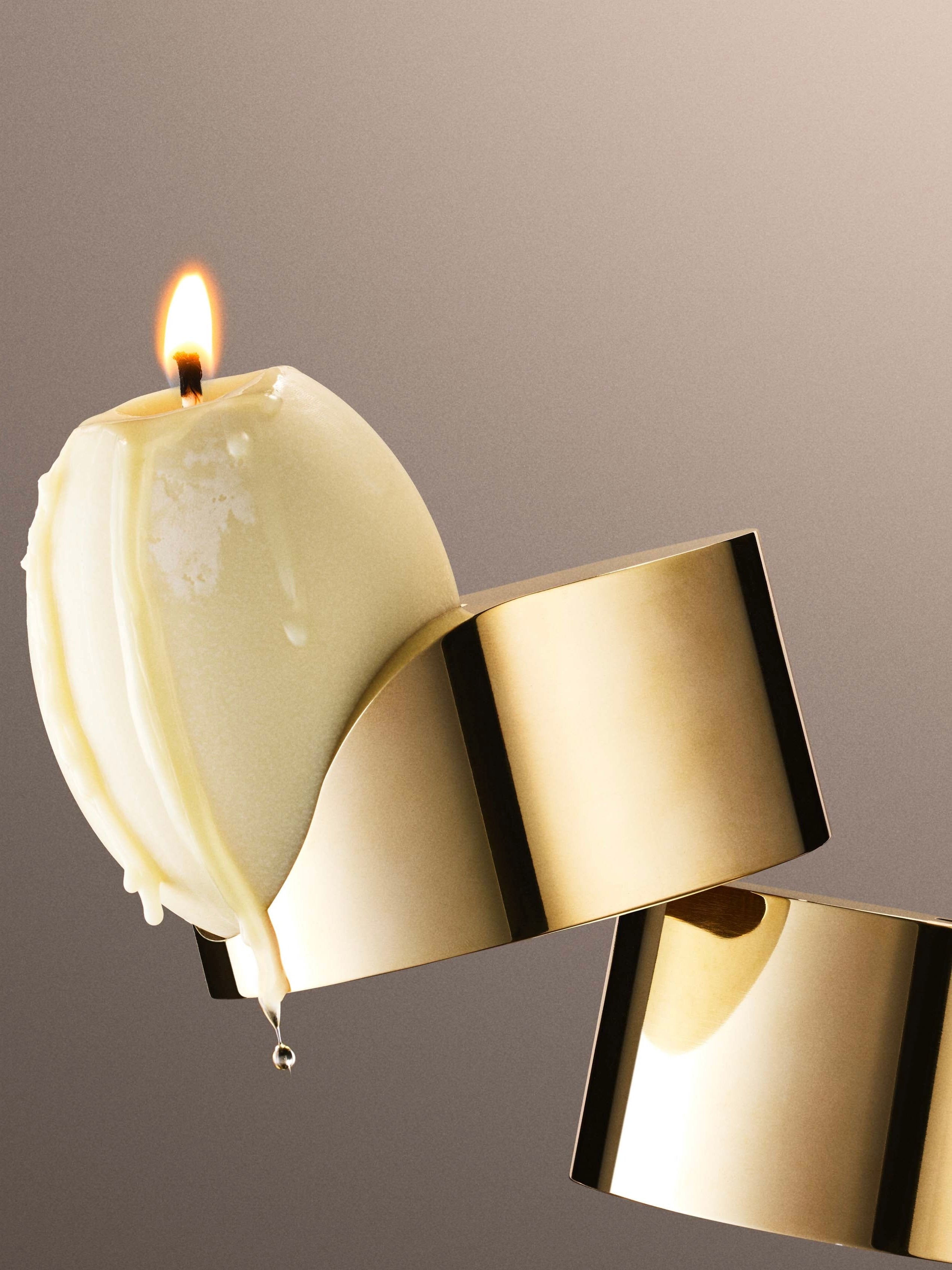 A lit round candle with melting wax sits atop a Low Perch Candle Holder by Tête Studio, its reflective gold surface gently curving. The soft flame casts a gentle light, and wax drips elegantly down the sides of the candle. The background smoothly transitions from beige to brown, emphasizing the candle's glow.