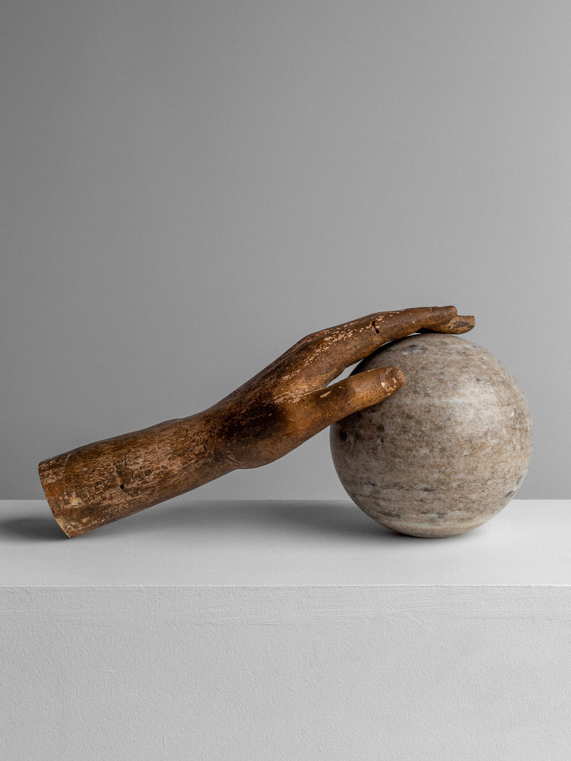 The Spigel Antique Wooden Hand Sculpture serves as a decorative accent, gently resting on a smooth, round stone set against a simple gray background. This minimalist scene highlights the detailed craftsmanship of the textured wood contrasted with the polished surface of the stone.