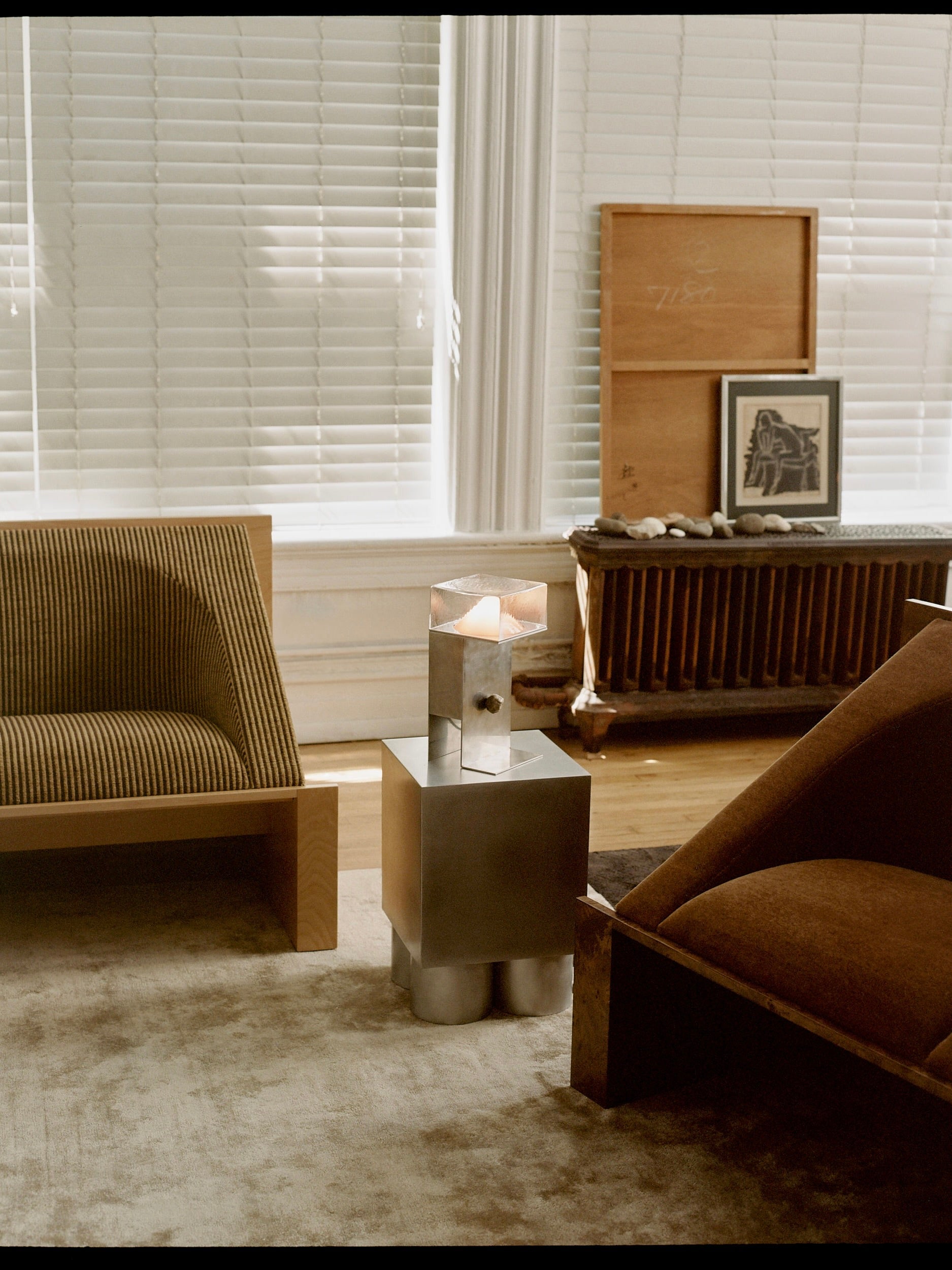 A cozy living room embodies minimalist values with the 'Perceptions' Sofa Bench by Known Work, two modern chairs, a metal side table holding a candle, and a framed picture against tall blinds. Warm light filters through, casting gentle shadows on the textured rug and enhancing serenity.