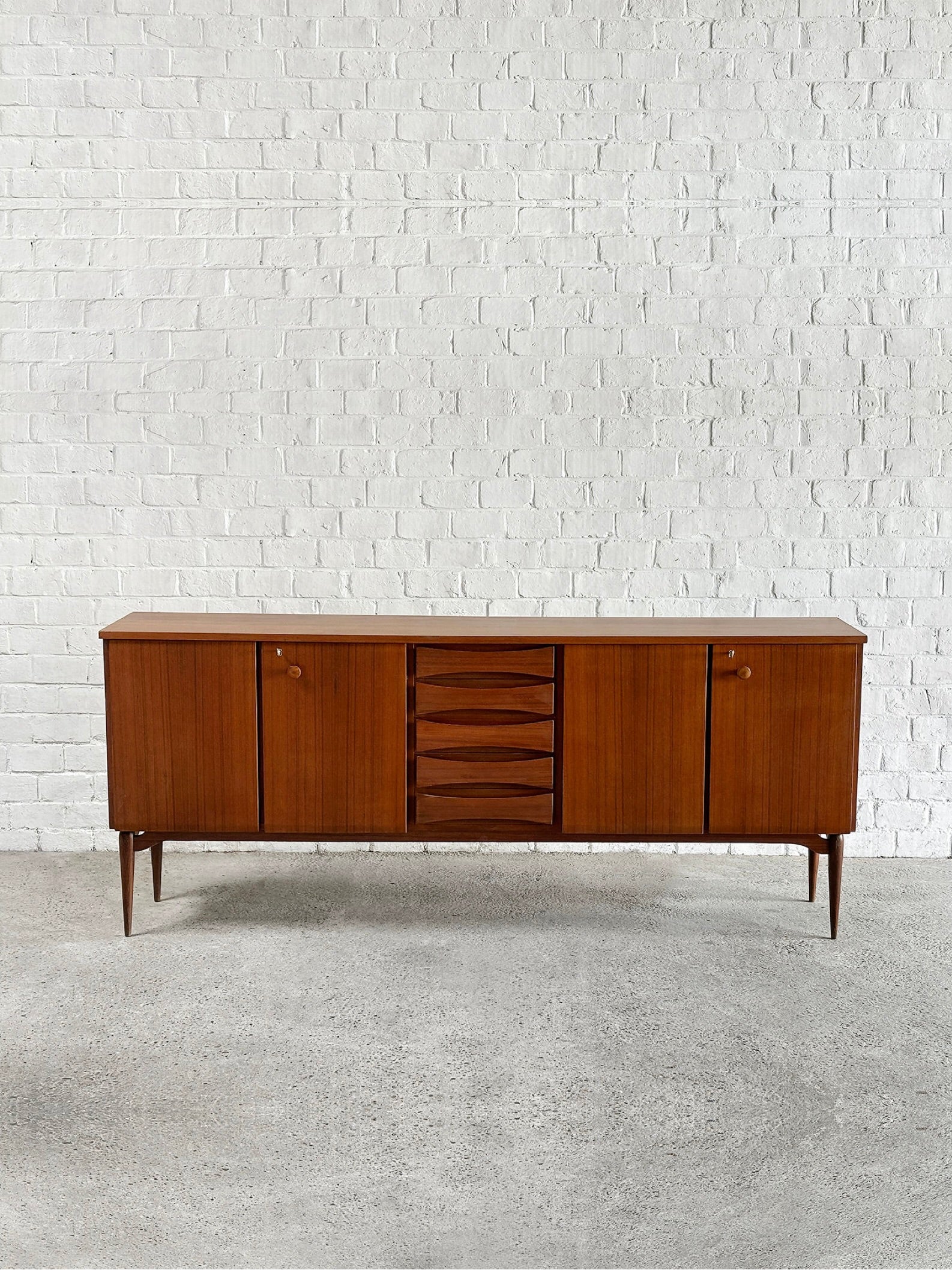 The Introverso Italian Modernist Teak Sideboard by Vittorio Dassi from the 1960s features four cabinet doors and three central drawers. This mid-century piece stands on tapered legs against a textured white brick wall on a concrete floor.