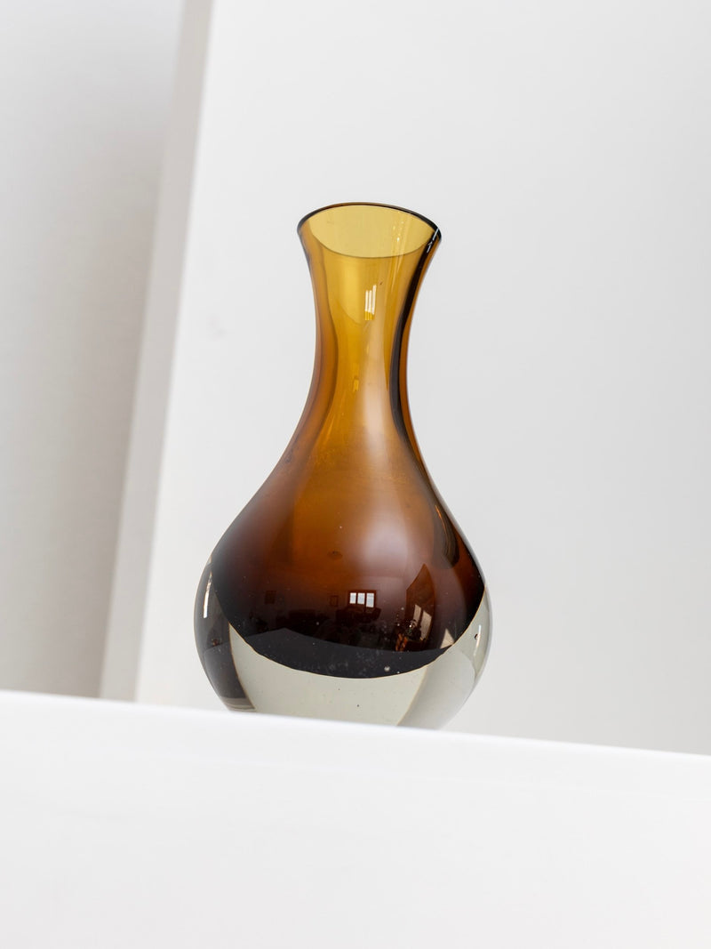 A vintage glass vase from Bottega Jacobs for Riihimäen Lasi Oy, featuring a mouth-blown brown design with a narrow neck and wide base, is set against a white background, exuding a minimalist and elegant style reminiscent of Tamara Aladin’s iconic work.