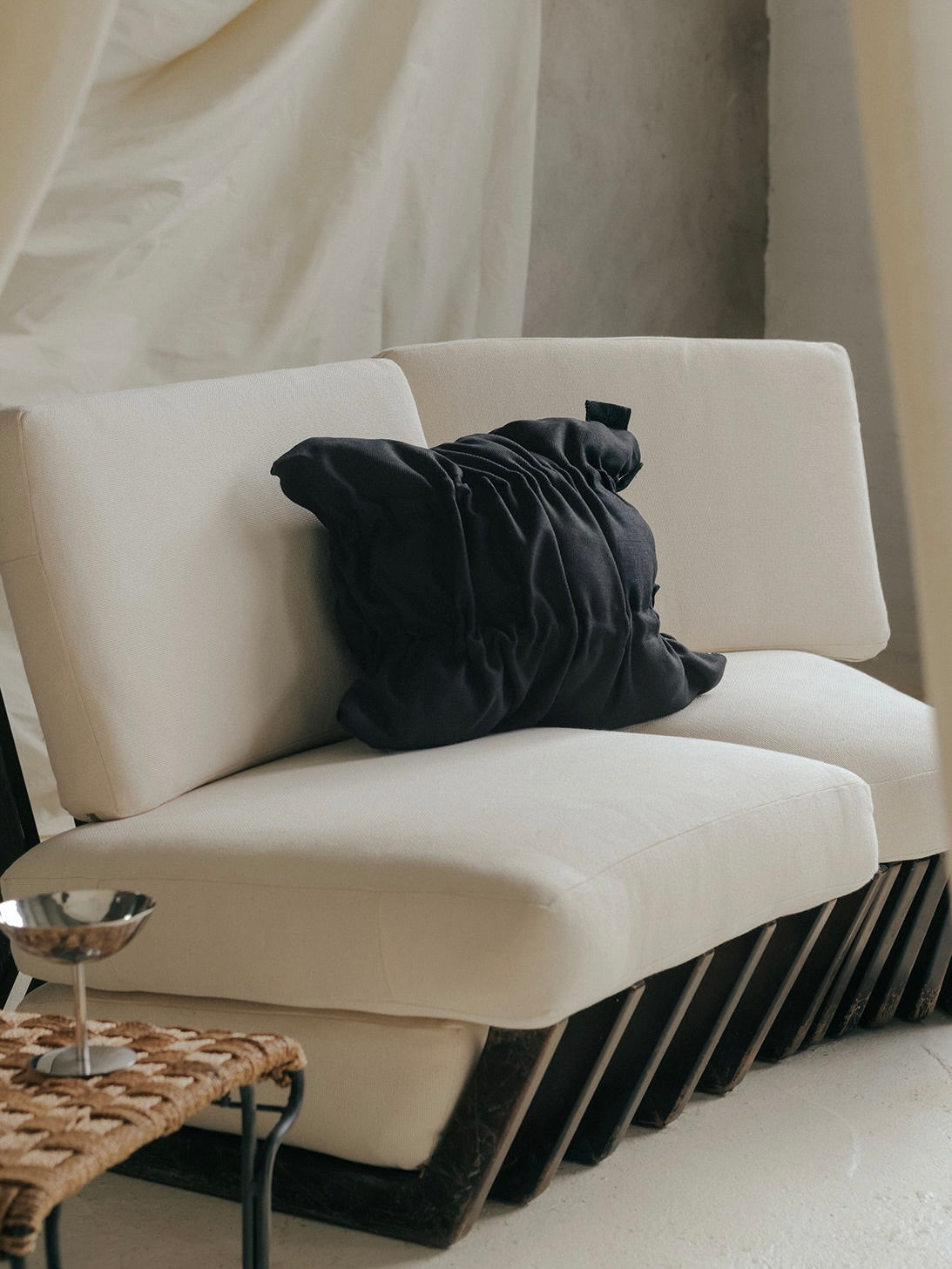 A modern sofa with beige cushions features the Daphne Charcoal Cushion by SANTA LIVING. A woven stool—handmade in Barcelona—supports a small silver cup in the foreground, enhancing a cozy setting against a light wall and showcasing exquisite home textiles.