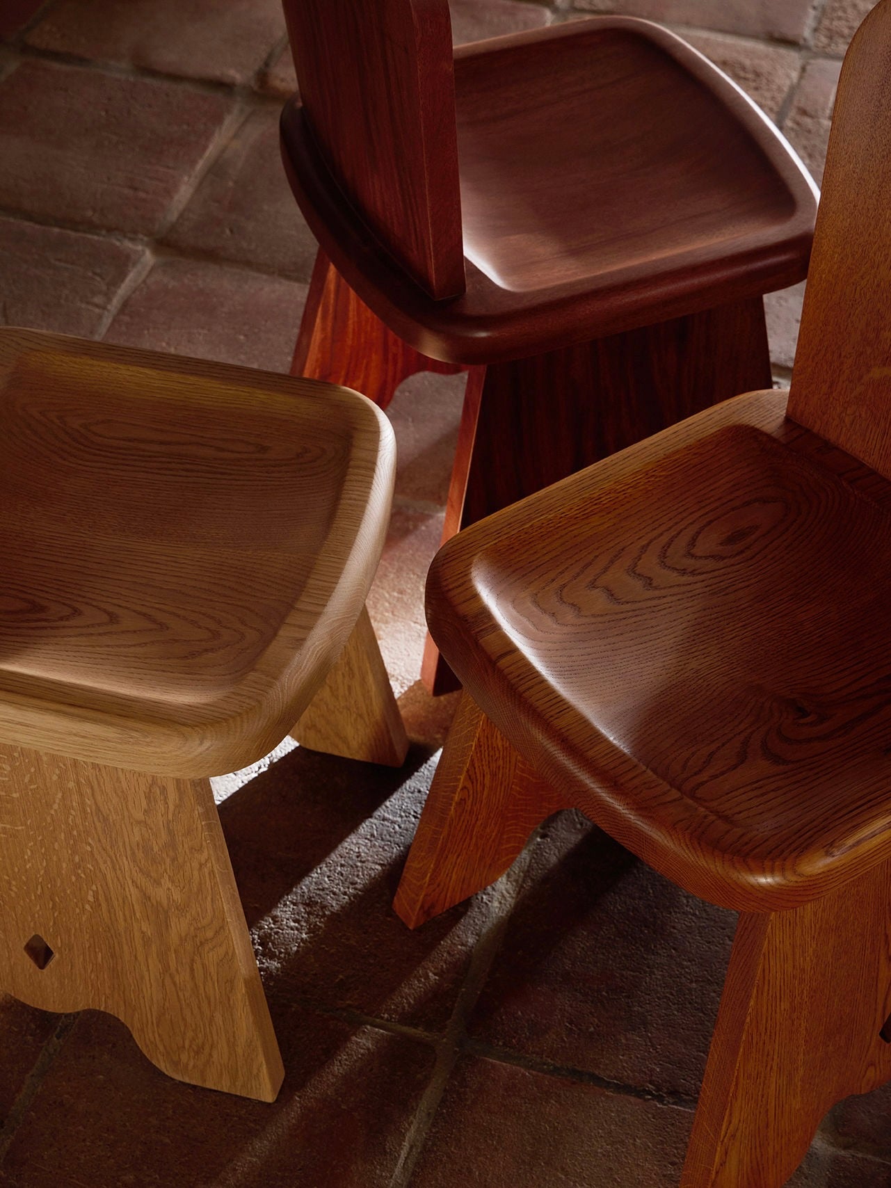 Three Lemon Rambling Chairs in African Mahogany Wood, each showcasing a unique wood grain from light to dark brown, with smooth curved seats, are arranged closely together on a tiled floor, highlighting their craftsmanship and contemporary elegance.